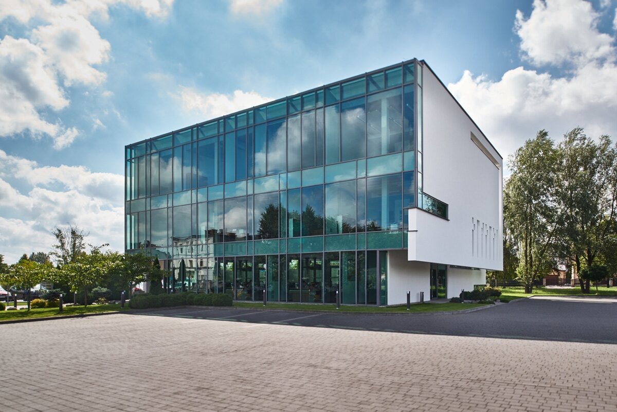 Na zdjęciu przedstawiona jest siedziba firmy Optopol z Zawiercia. Budynek jest w kształcie prostokątnej bryły. Jej ściana frontowa jest przeszklona, ściana boczna zbudowana jest z muru koloru białego. Podłoże przed siedzibą firmy jest wyłożone kostką brukową.