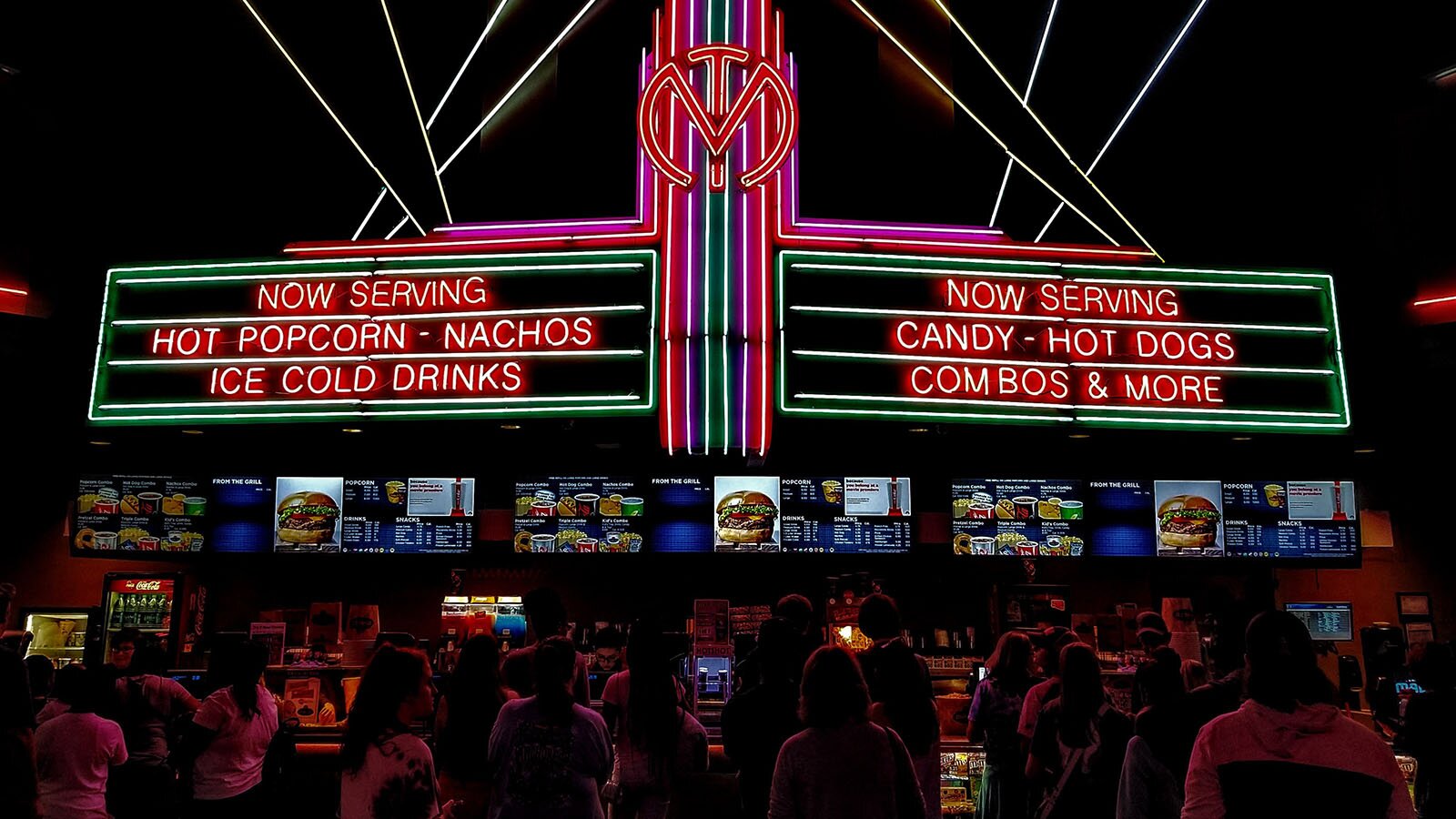 Zdjęcie ukazuje tłum ludzi stojących przed kontuarem w barze szybkiej obsługi. Nad nimi znajduje się menu i pokazane są dostępne dania, takie jak hamburgery czy napoje. Na samej górze znajdują się neonowe napisy po angielsku Now serving, Hot Popcorn, nachos, ice cold drinks, candy, hot dogs, combos and more.