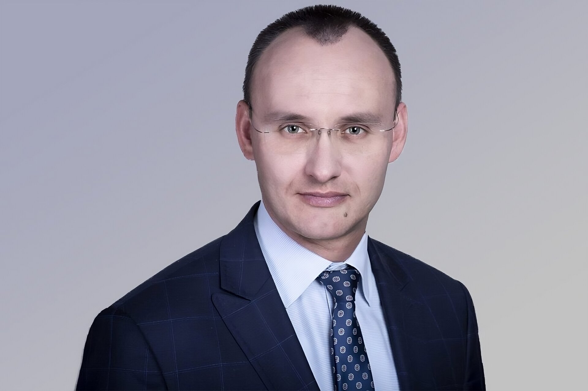 Na zdjęciu mężczyzna w wieku średnim. Z bardzo krótką fryzurą. Mocno widoczne zakola, przedzielone fragmentem włosów. Lekko uśmiechnięta twarz. Nosi okulary. Ubrany w garnitur. Pod garniturem widoczna koszula z krawatem.