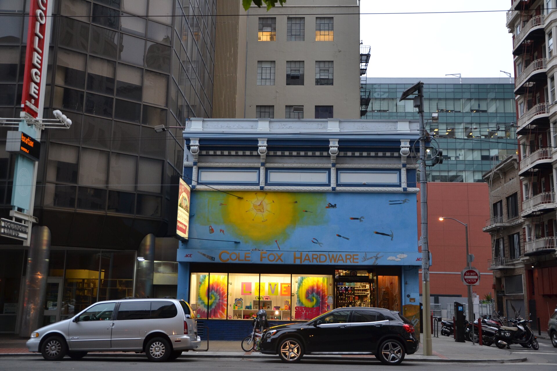 Fotografia przedstawia fragment gęstej zabudowy San Francisco. Na pierwszym planie znajduje się jezdnia z zaparkowanymi samochodami, za nimi witryna oraz wieżowce, jeden przy drugim.