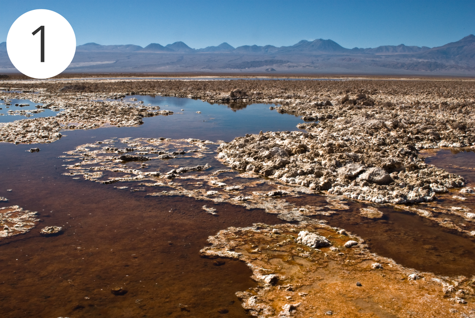 Zdjęcie przedstawia fragment pustyni Atacama na terenie Chile. Znajdują się tam szaro—brązowe złoża saletry chilijskiej poprzecinane małymi zbiornikami wodnymi. W tle są góry, a nad nimi niebieskie niebo, które odbija się w dalej położonych zbiornikach wodnych.