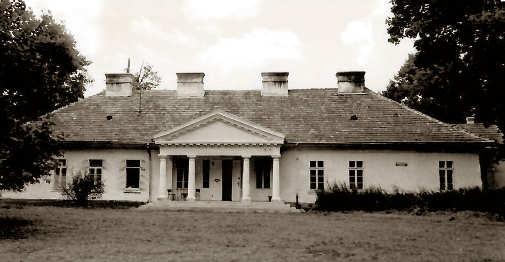 Czarno‑biała fotografia przedstawia uszkodzony dworek. W budynku nie ma niektórych okien, jedno skrzydło drzwi jest urwane. Dworek posiada cztery kominy oraz wejście główne zadaszone portykiem.  Obok dworku rosną drzewa oraz krzewy. 