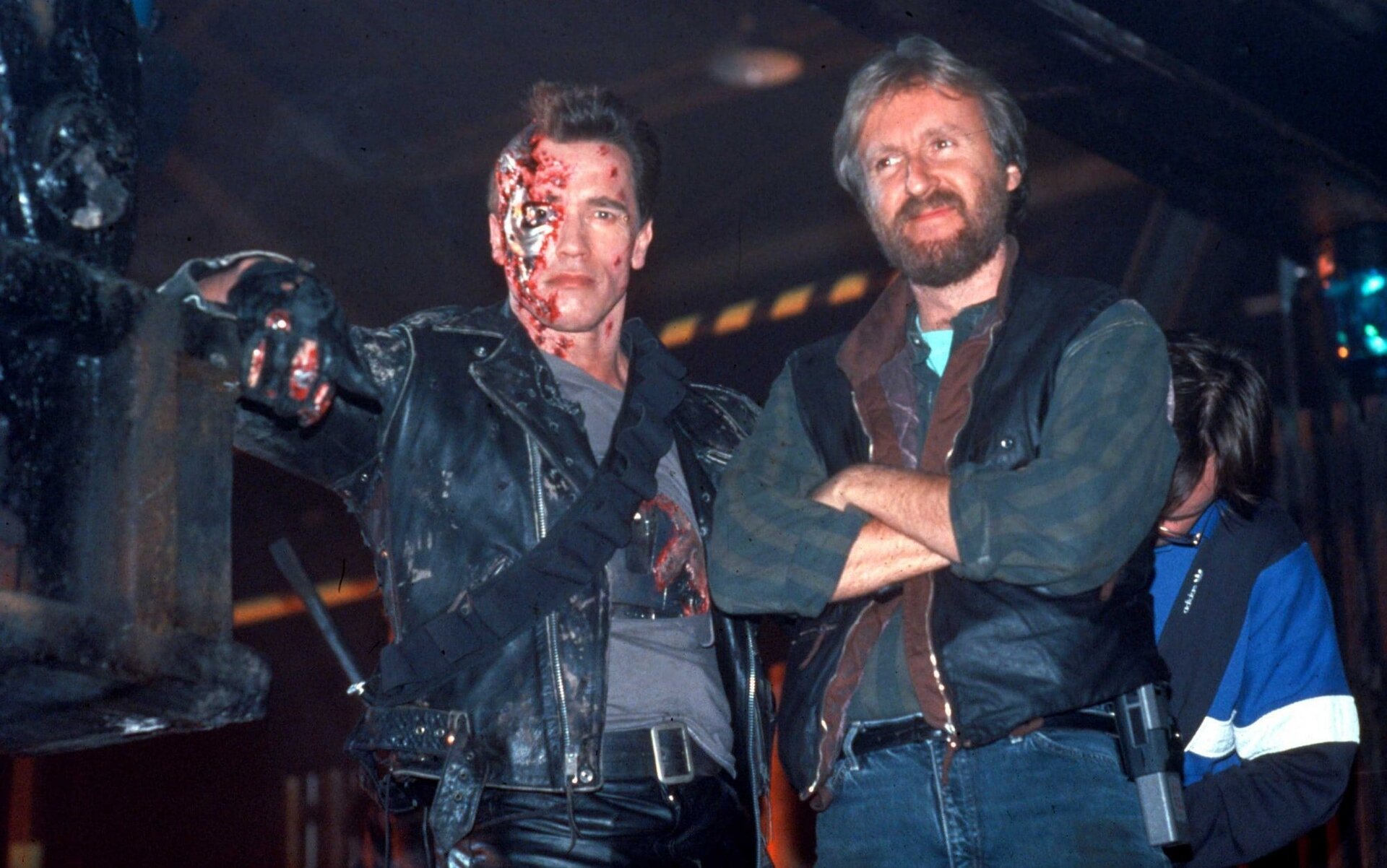 Fotografia przedstawia aktora, którym jest Arnold Schwarzenegger i reżysera Jamesa Camerona na planie filmu „Terminator 3”. Schwarzenegger stoi po lewej stronie i został sfotografowany jako mężczyzna w średnim wieku, ubrany w ciemne skórzane spodnie i skórzaną kurtkę, spod której widoczna jest szara koszulka z czarno‑czerwoną grafiką. Na jego twarzy widoczna jest charakteryzacja filmowa – liczne rany i popękana cyberskóra, wykonana głównie na prawej stronie jego twarzy. Po prawej stronie stoi reżyser filmu, James Cameron - mężczyzna w średnim wieku o średniej długości ciemnym zaroście i siwych włosach. Cameron sprawia wrażenie zadowolonego, jest uśmiechnięty, a jego spojrzenie skierowane jest w bok. Ma na sobie czarno‑brązową kamizelkę, zieloną koszulę w paski oraz niebieskie jeansy. Jego ręce są skrzyżowane. 