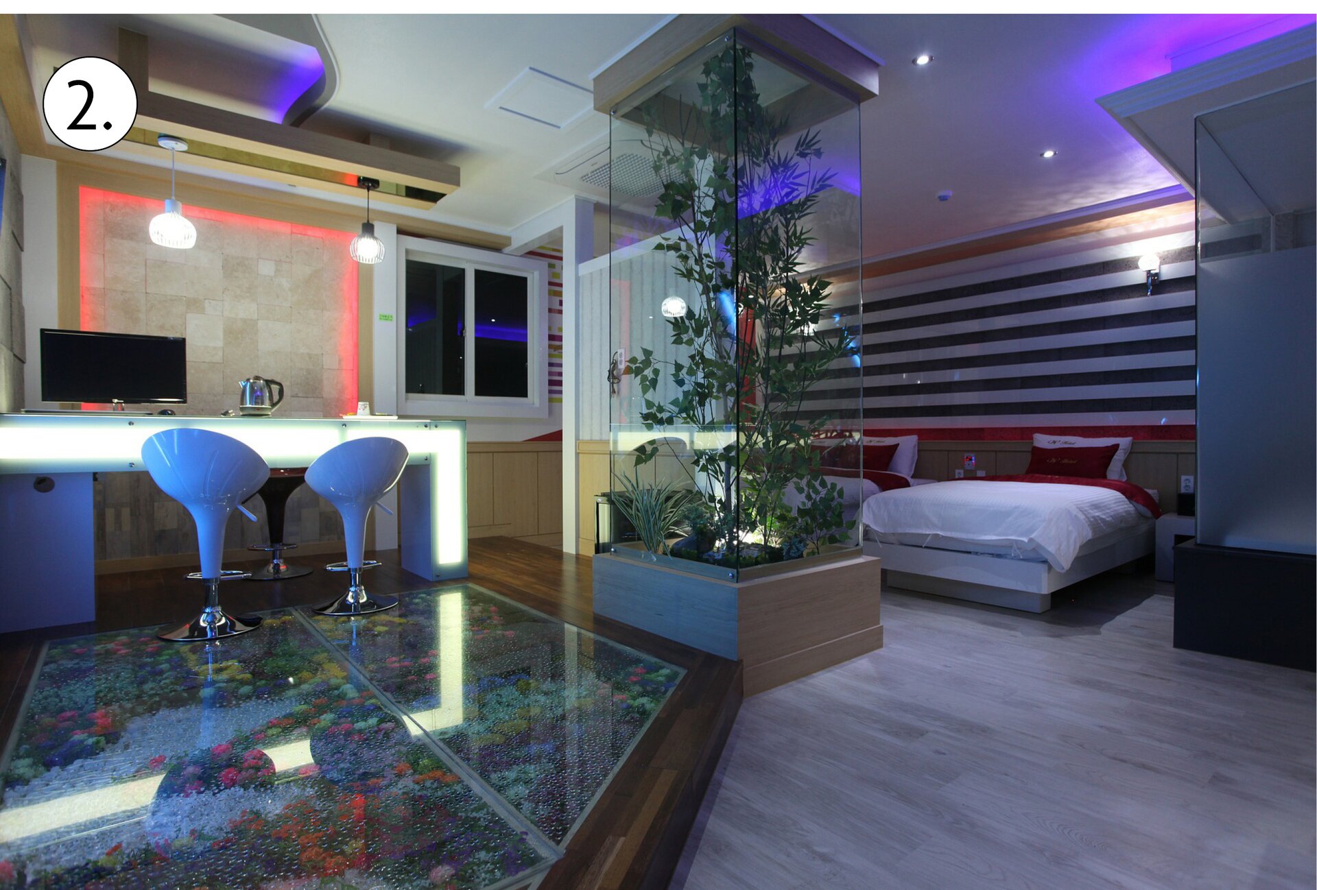 Ilustracja ukazuje nowoczesne wnętrze mieszkalne. Po lewej stronie widoczny jest blat z dwoma białymi fotelami. Na blacie znajduje się ekran oraz czajnik elektryczny. Podłoga w tym aneksie jest szklana, pod nią widoczna jest kolorowa kompozycja w donicy. Część sypialna z podwójnym łóżkiem jest oddzielona kwadratową donicą, obudowaną szklanymi ścianami. Wewnątrz niej znajduje się kompozycja kwiatowa.