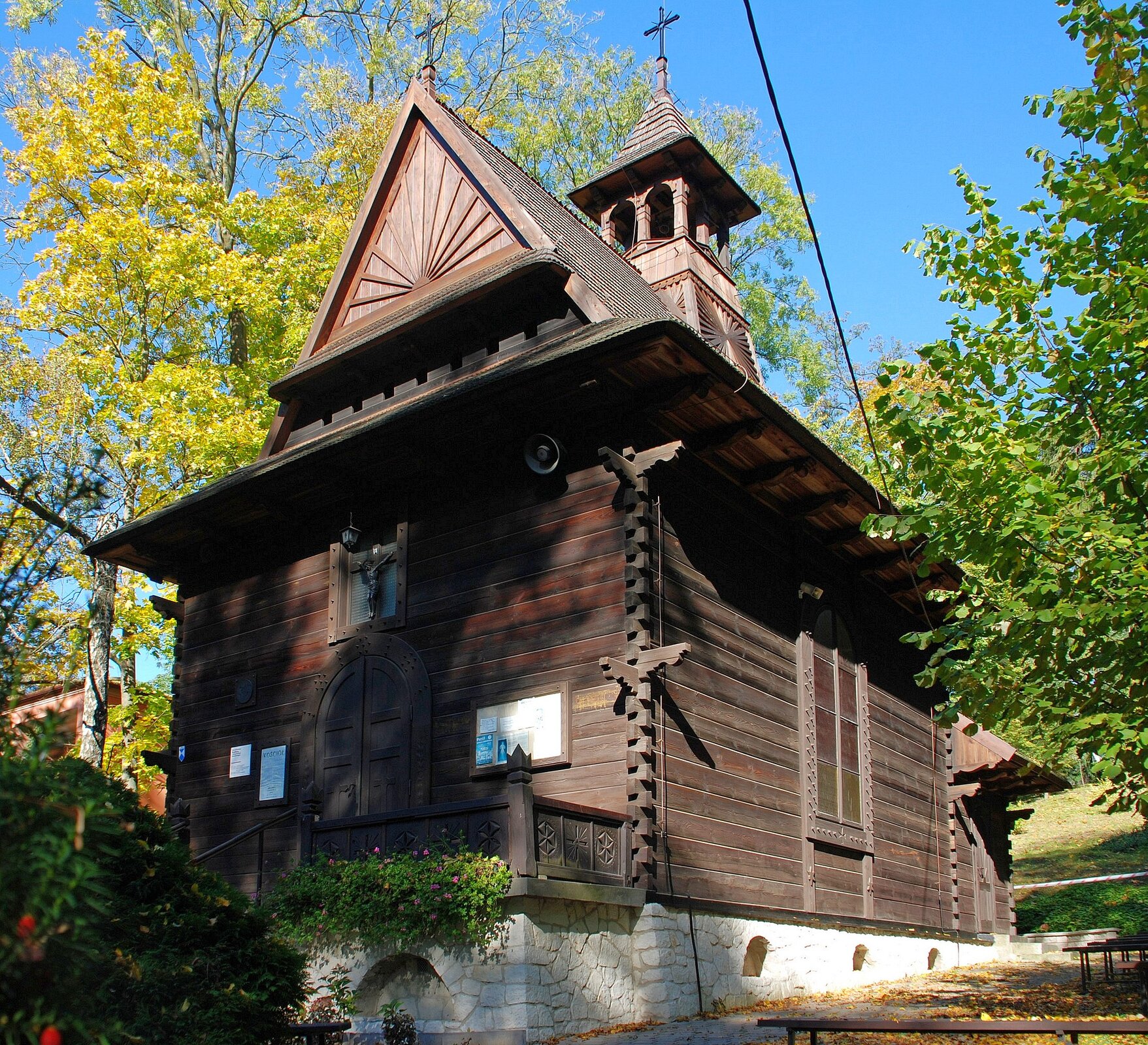 Na zdjęciu tradycyjny mały, drewniany kościół w lesie. Kościół znajduje się na białym fundamencie, ma jedną wieżę i dwa krzyże na dachu. Dookoła kościoła wysokie drzewa. 
