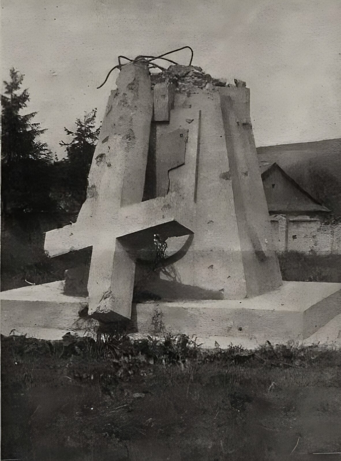 Zdjęcie przedstawia kamienny monument w kształcie piramidy z umieszczonym na nim krzyżem, który został uszkodzony pociskiem artyleryjskim i zwalony na ziemię. Widoczne są wychodzące z postumentu druty zbrojeniowe. W tle murowane ogrodzenie, drzewa, zabudowania.