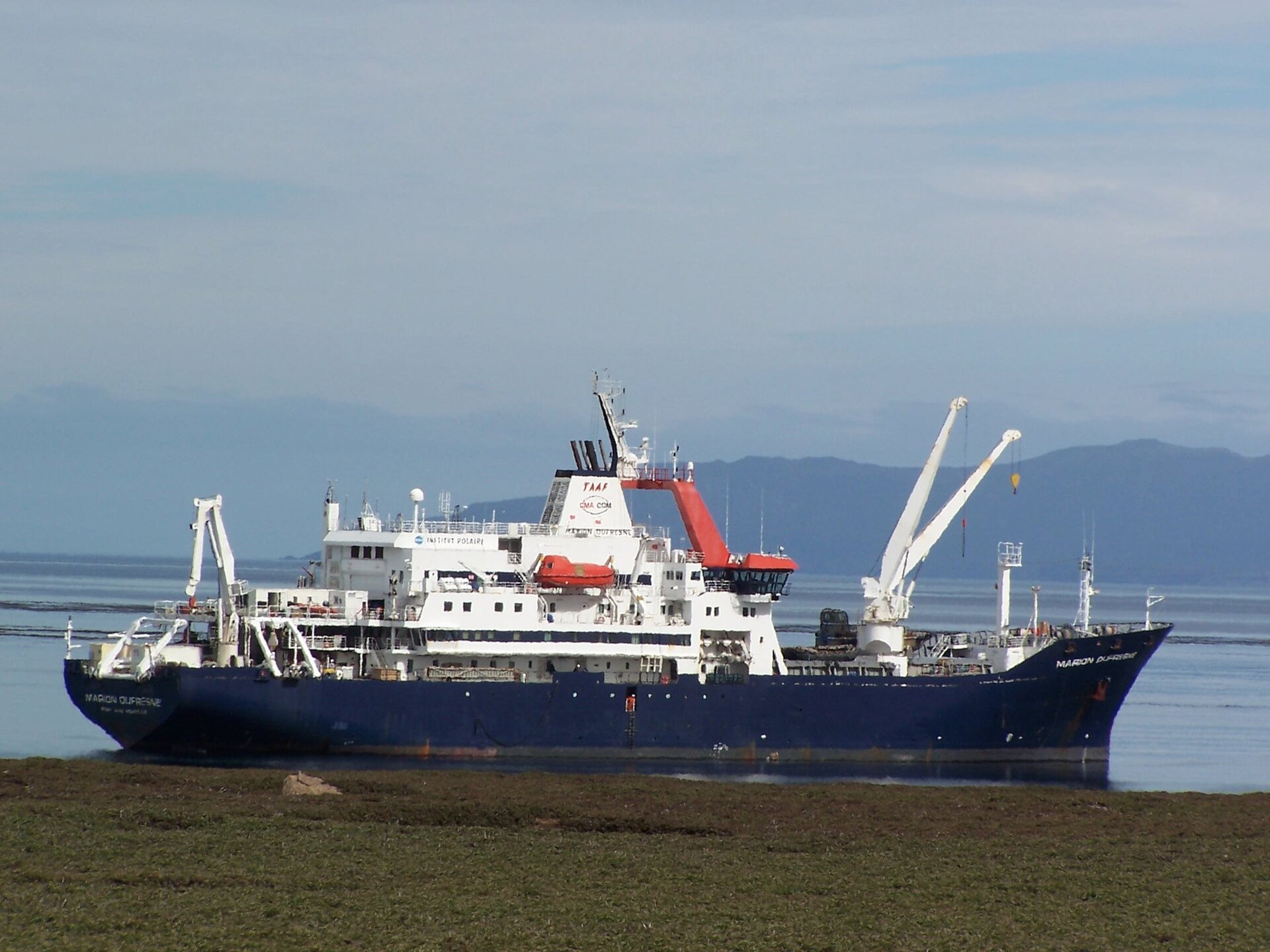 Zdjęcie przedstawia duży statek transportowy. Na pokładzie ma dźwigi. W tle jest pasmo górskie. 