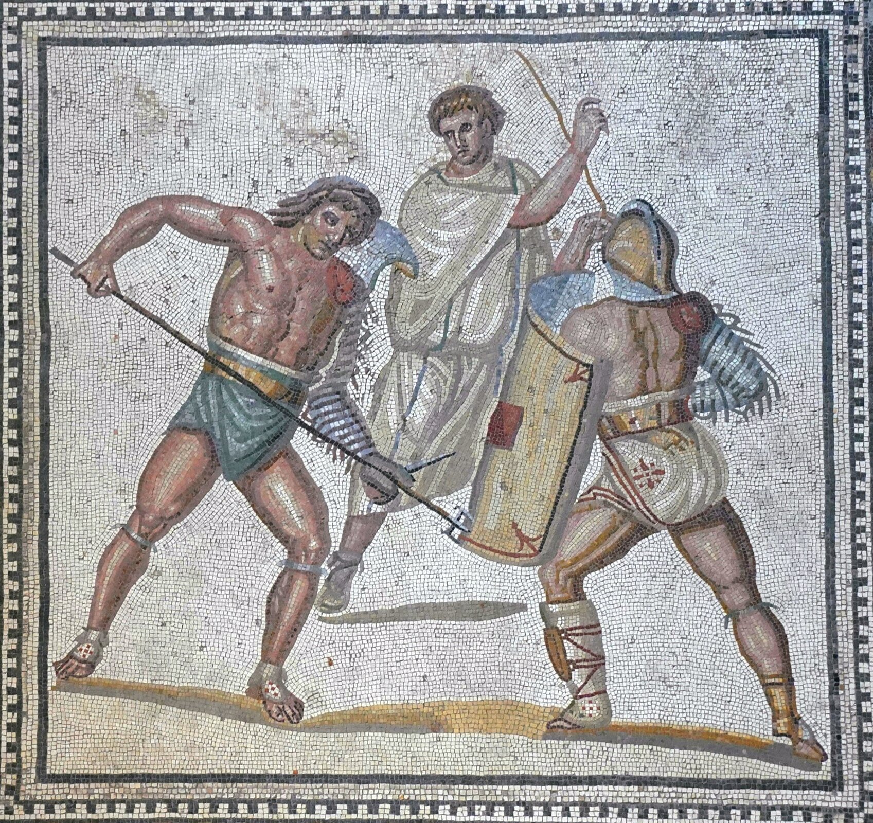 Mozaika przedstawiająca walczących gladiatorów. W kwadratowej ramce, na jasnokremowym tle ukazano trzech mężczyzn. Jeden z nich to młody, umięśniony mężczyzna trzymający w dłoni trójząb, którym celuje w nogę mężczyzny w hełmie z tarczą u boku, ukazanego tyłem. Na jego lewej nodze obecny jest ochraniacz. Za nimi obecny jest jasnowłosy mężczyzna w szacie sięgającej połowy łydki, z uniesioną prawą dłonią, patrzący w prawo. 