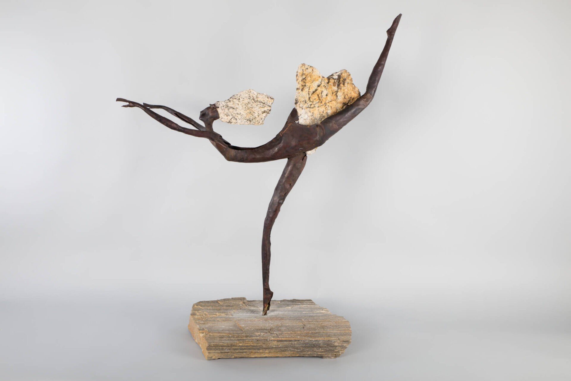 Fotografia przedstawia dzieło na tle szarej ściany. Podstawę stanowi nierówna drewniana bryła, na której umieszczono postać stojącą na jednej nodze. Jej druga noga wyciągnięta jest do tyłu oraz ku górze, co sprawia wrażenie, że jest baletnicą. Długie ręce postaci wyciągnięte są przed siebie, jej tułów jest więc prostopadły do podłoża, a plecy wygięte w łuk. Włosy postaci wykonano z beżowo‑żółtego materiału, podobny element został użyty jako spódniczka. Jej kończyny są długie oraz smukłe, a ciało wykonane z ciemnego metalu.