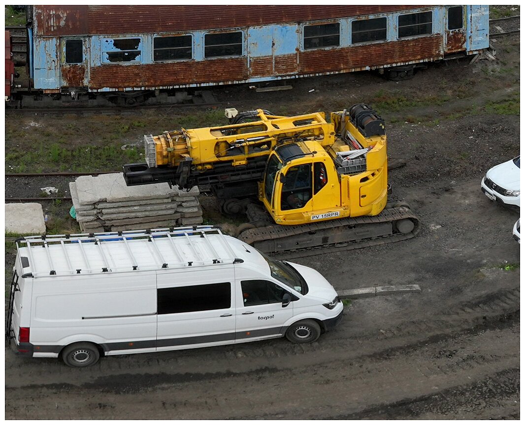 Ilustracja przedstawia palownicę w trakcie pracy na budowie. Maszyna na zdjęciu posiada podwozie gąsiennicowe, ramię oraz urządzenie wiertnicze.