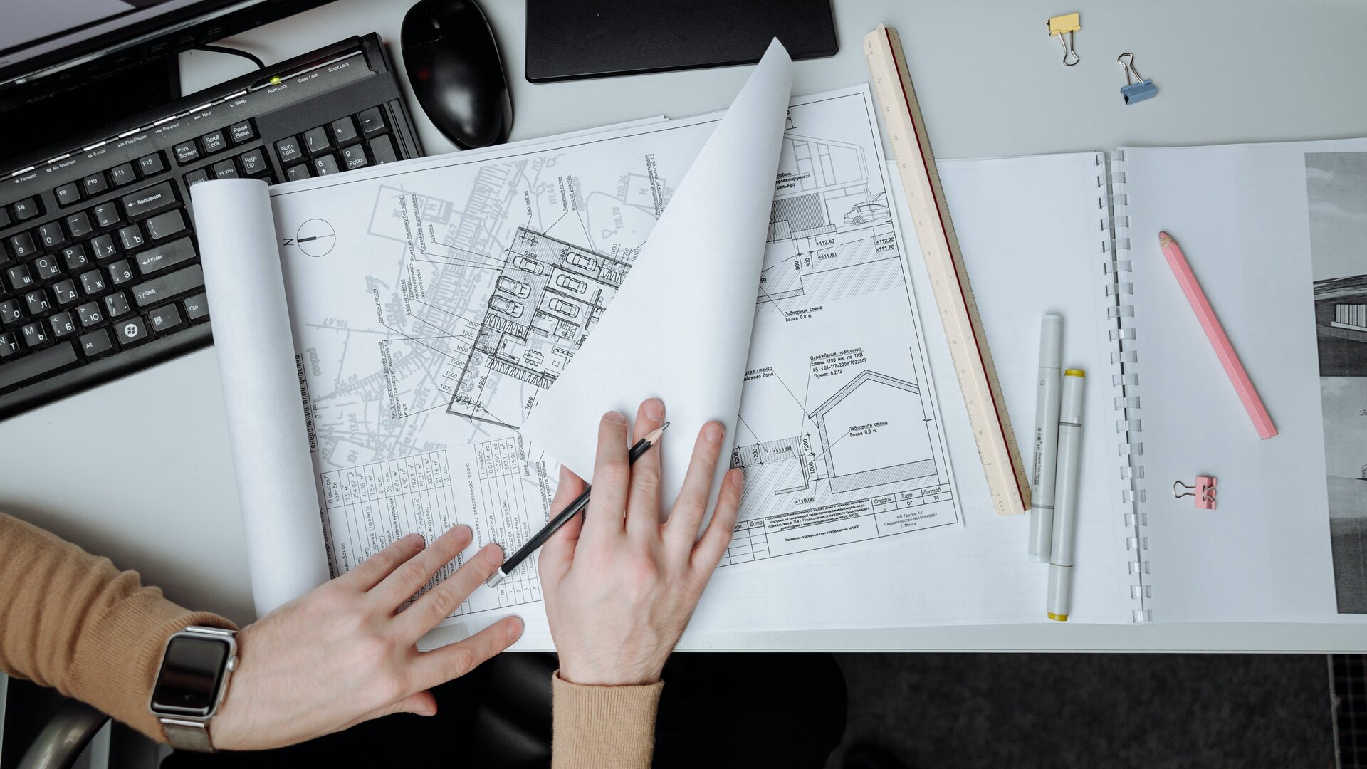 Ilustracja przedstawia biurko, na którym leżą projekty architektoniczne rozrysowane na kartkach. Kartki przewracane są przez osobę będącą przy biurku. Postać nie jest w całości widoczna na ilustracji. Widoczne są tylko jej ręce. Lewą dłoń opiera na projektach. Prawą przewraca strony. Oprócz planów architektonicznych na biurku znajdują się także: monitor, klawiatura i myszka oraz zeszyt i długopisy. 