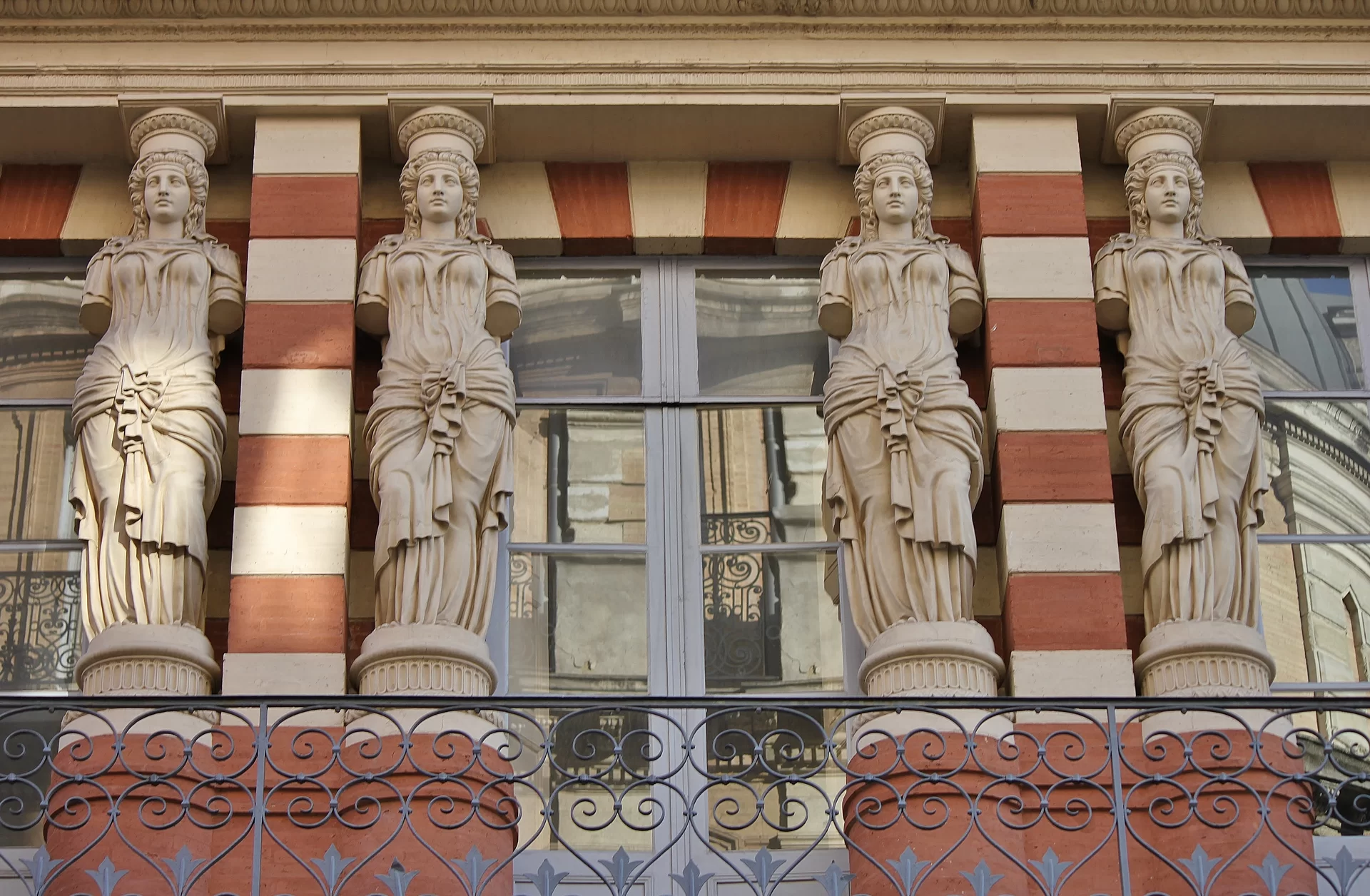 Zdjęcie przedstawia fragment budynku z zewnątrz. Na jednym z poziomów ustawiono cztery kolumny z jasnego kamienia w kształcie stojących kobiet w długich szatach. Ustawione są następująco: rzeźba, kwadratowa kolumna, rzeźba, okno i znów rzeźba, kwadratowa kolumna, rzeźba. Kwadratowe kolumny mają biało‑pomarańczowe pasy.