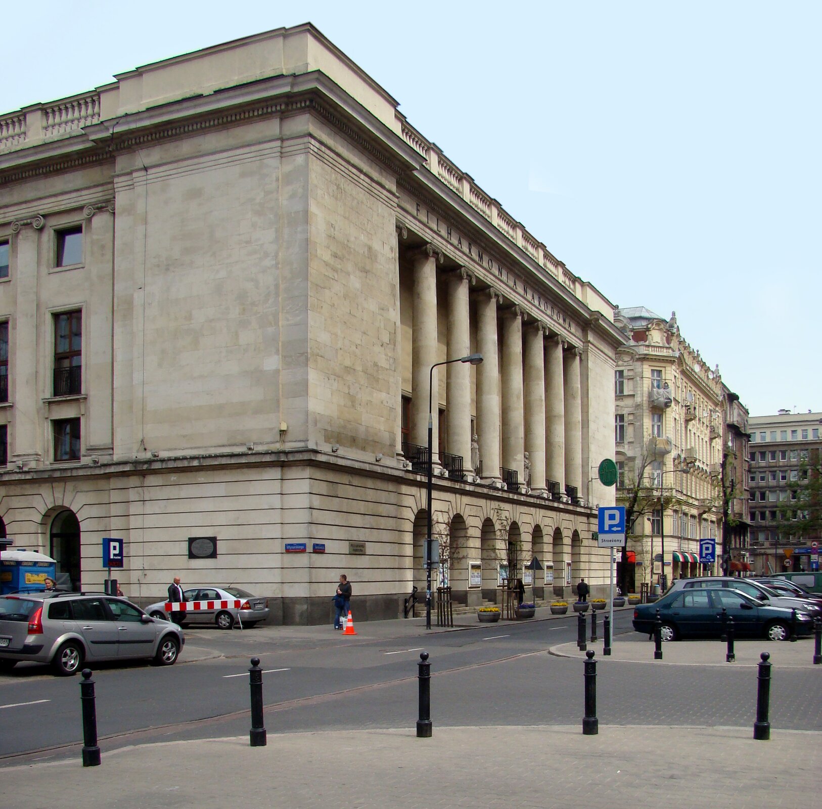 Ilustracja przedstawia budynek Filharmonii Narodowej w Warszawie, gdzie odbywa się Międzynarodowy Konkurs Pianistyczny im. Fryderyka Chopina. Budynek jest duży, czteropiętrowy, w stylu klasycystycznym. Ma arkady i kolumny, wybudowany jest z jasnego kamienia.