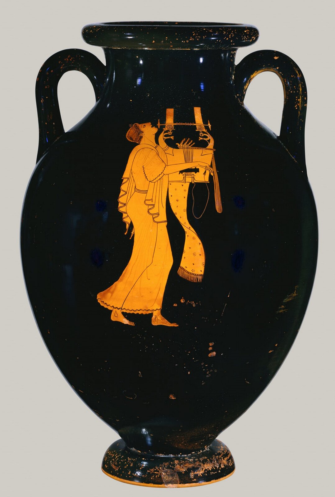  Ilustracja przedstawia wazę „Amphora przedstawiająca poetę”. Na wazie widnieje malowidło postaci. Jest to mężczyzna, ubrany w długi szaty. Postać idzie przed siebie, a w rękach trzyma instrument muzyczny, na którym gra.