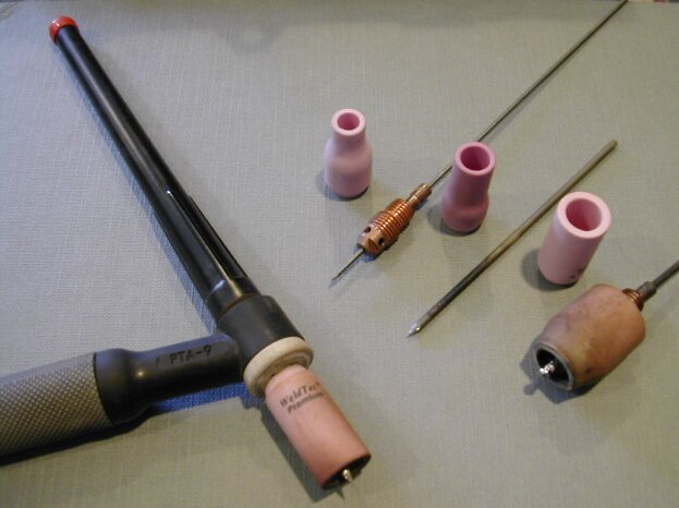 Zdjęcie przedstawia uchwyt do spawania metodą TIG. Ma on kształt litery L z wydłużonym jednym przy zakrzywieniu. Obok uchwytu widoczne są różnorodne końcówki. Różnią się one grubością.