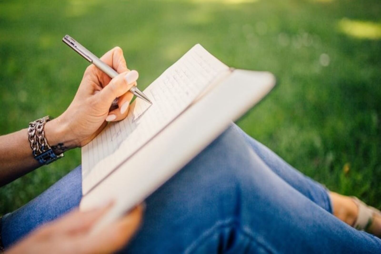 Zdjęcie pokazuje trzymany na kolanach zeszyt oraz kobiecą rękę z długopisem, piszącą w tym zeszycie. Fotografia obrazuje robienie notatek z wywiadu.