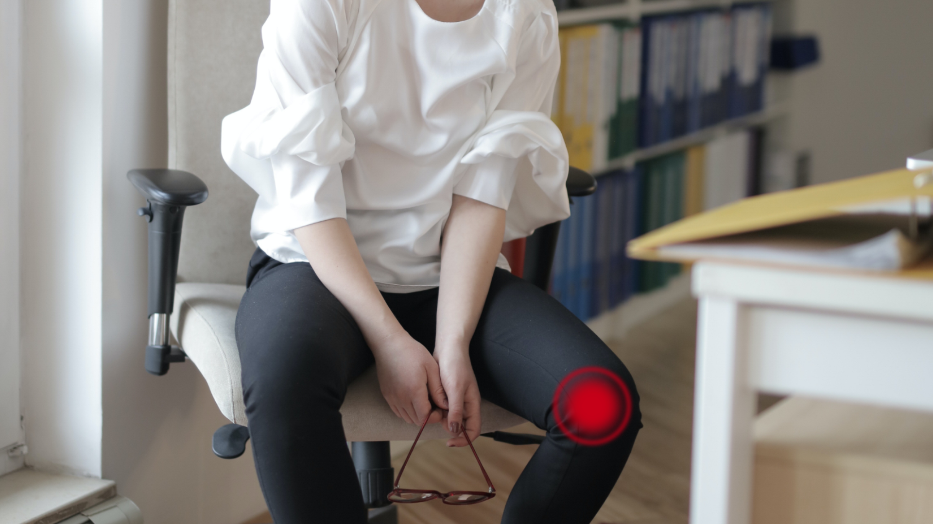 Zdjęcie przedstawia kobietę siedzącą na krześle biurowym. Nie widać twarzy kobiety. Na jednym kolanie znajduje się czerwony punkt przedstawiający źródło bólu.
