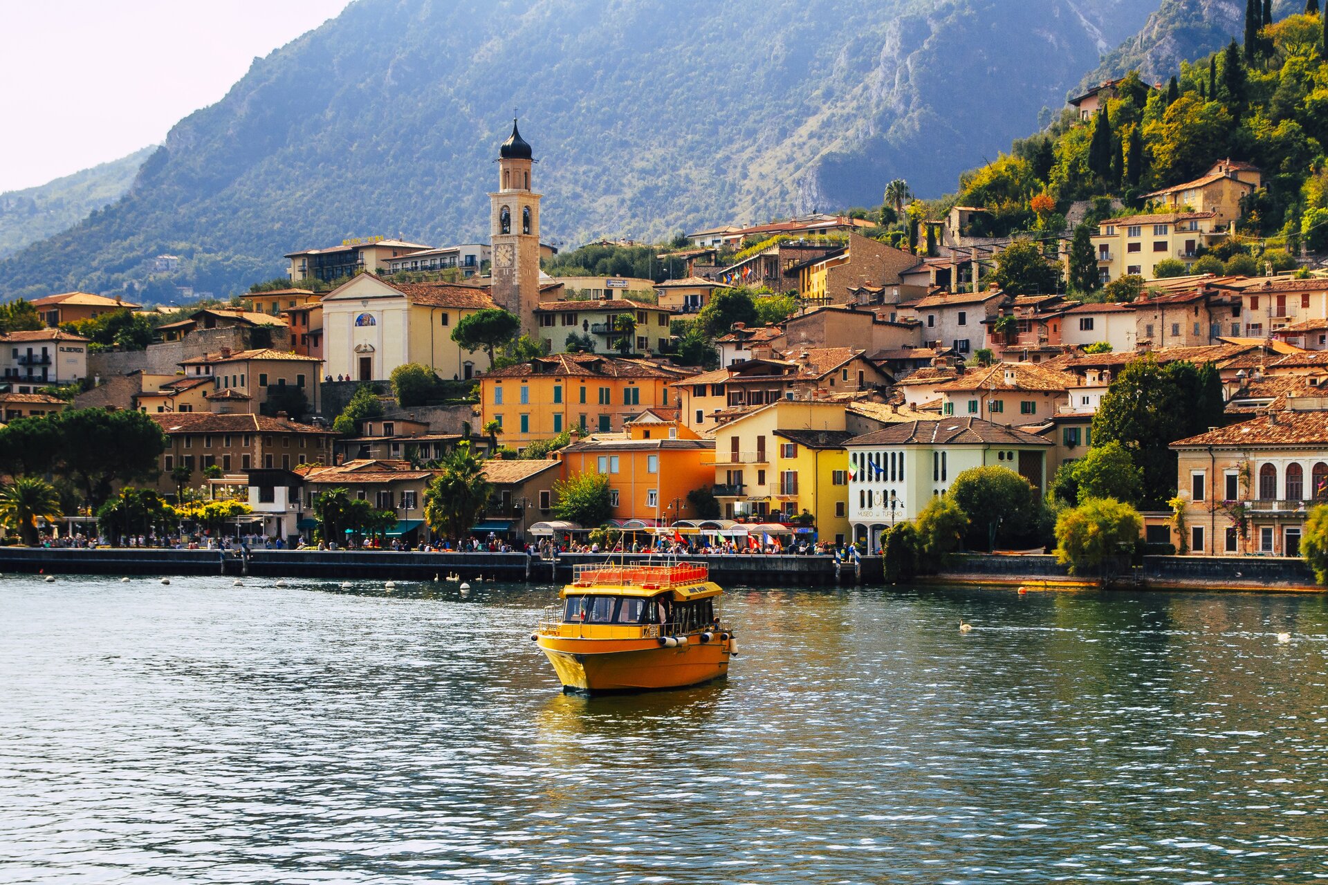 Ilustracja przedstawia jezioro otoczone górami. Pomiędzy jeziorem, a górami rozpościera się miasteczko, brzegiem jeziora spacerują ludzie. Po jeziorze pływa łódka.