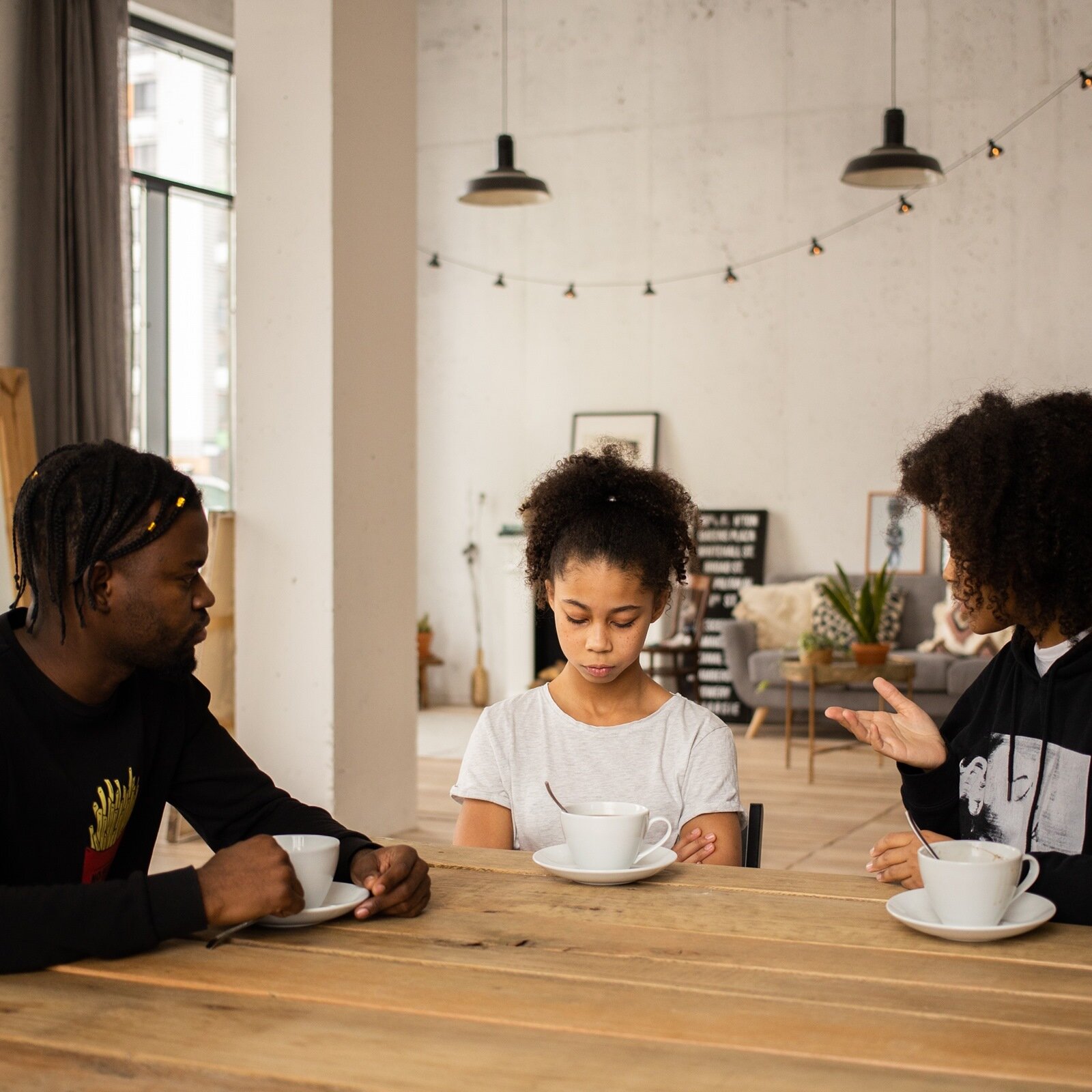 Zdjęcie przedstawia trzy osoby siedzące przy drewnianym stole w salonie. Po lewej stronie widoczny jest mężczyzna, zwrócony w prawo, który w prawej ręce trzyma uszko filiżanki stojącej przed nim. Pośrodku widoczna jest nastolatka, która ma spuszczoną głowę i założone na brzuchu ręce. Przed nią stoi filiżanka, w której zanurzona jest łyżeczka. Po prawej stronie widoczna jest kobieta, zwrócona w lewo, w stronę nastolatki, która gestykuluje. W tle widoczne jest wnętrze pokoju z wyposażeniem.