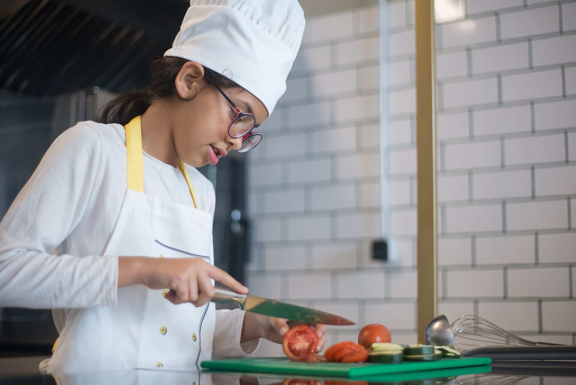 Zdjęcie przedstawia dziewczynkę stojącą przy blacie kuchennym. Dziewczynka ma na sobie fartuszek oraz nakrycie głowy kucharze. Długie ciemne włosy ma upięte z tyłu. Dziewczynka ma lekko ugięte ręce. W prawej ręce trzyma duży nóż kuchenny, którym kroi pomidora leżącego na zielonej desce do krojenia. Lewą ręką przytrzymuje krojony pomidor. Na desce do krojenia widoczne są cały pomidor, plastry pomidora i cukinii. Na prawo od deski widoczna jest chochelka i trzepaczka.
