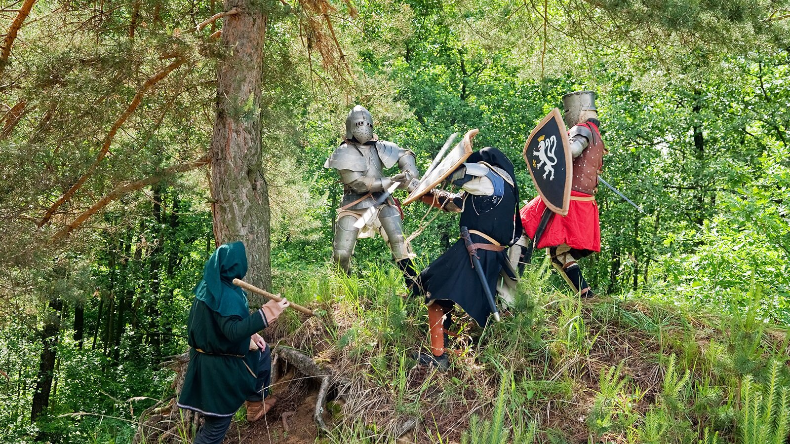 Zdjęcie przedstawia cztery osoby ubrane w średniowieczne stroje rycerskie i walczące między sobą w lesie. 
