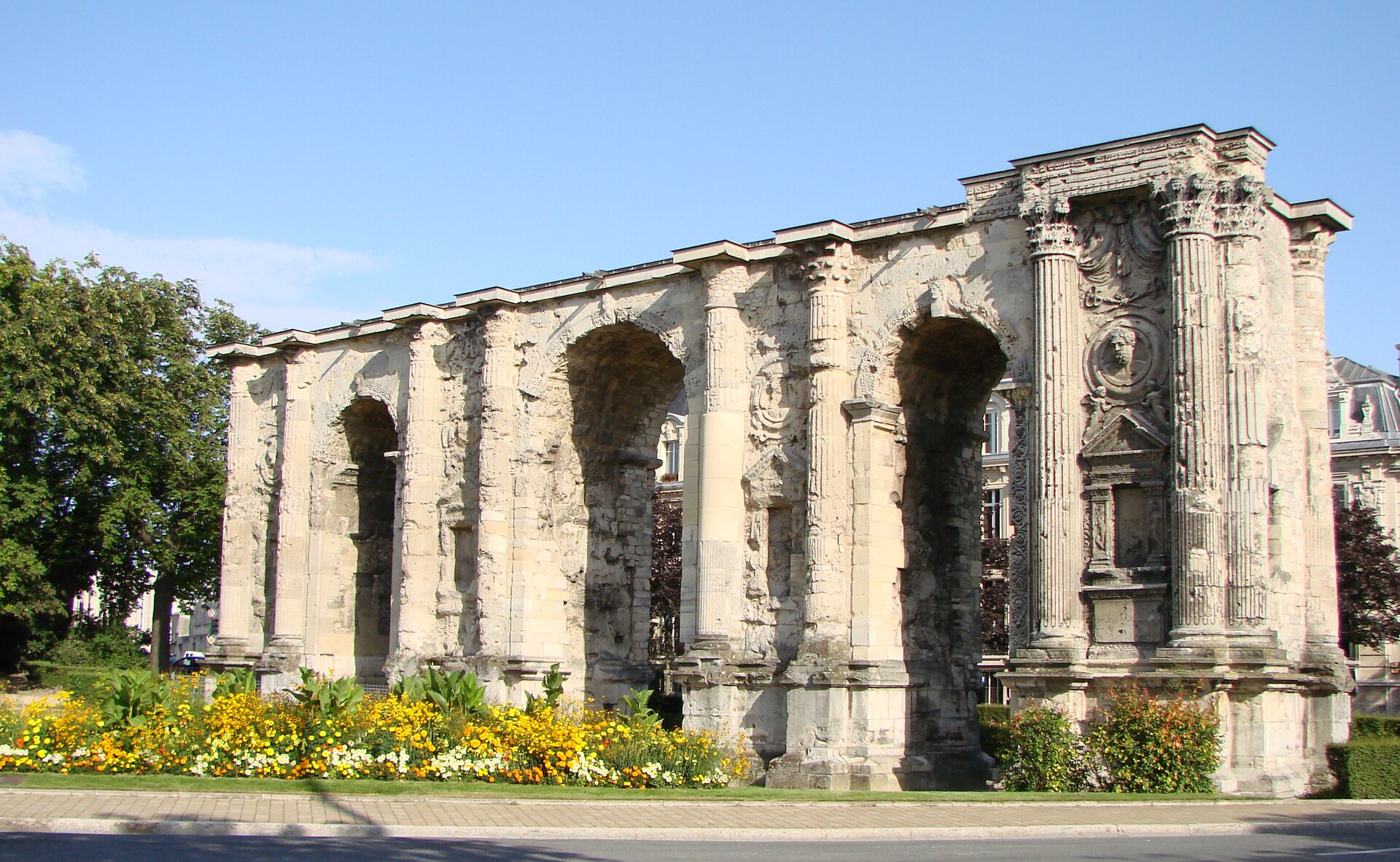 Fotografia przedstawiająca łuk triumfalny – jasną budowlę z trzema łukowymi otworami - przejściami. Między otworami znajdują się kolumny z podłużnymi żłobieniami oraz ozdobnymi fryzami, a także niewielkie rzeźby. Przed budynkiem rosną kwiaty, a dalej - drzewa.