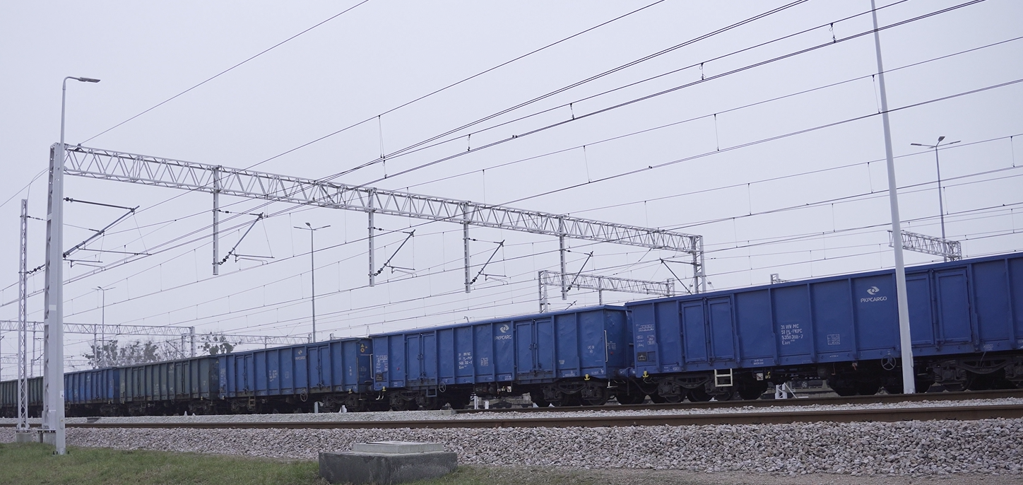 Zdjęcie pokazuje sieć trakcyjną. Widoczne są tory, po jednym z nich jedzie skład wagonów towarowych. Nad torami, na metalowej konstrukcji wsporczej rozwieszonej poziomo w poprzek torów, zawieszone są przewody trakcyjne.