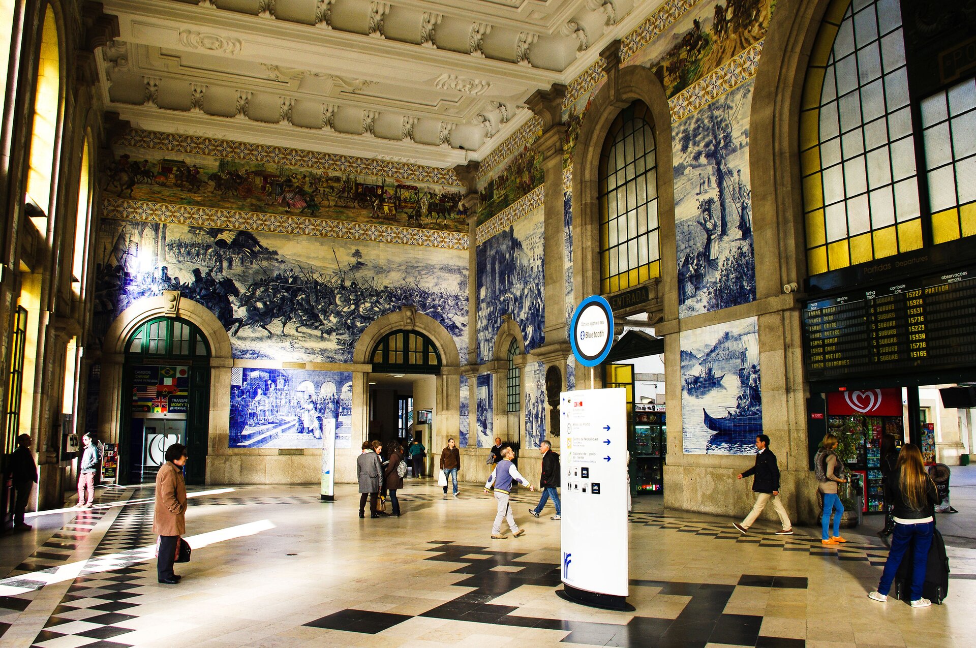 Zdjęcie przedstawia wnętrze Dworca Kolejowego w Porto w Portugalii. Podróżni idą w różnych kierunkach przez salę. Ściany wyłożone malowanymi płytkami ceramicznymi ukazują sceny historyczne wielkich rozmiarów, malowane  jednym kolorem o zimnej barwie. Nad nimi, pasem biegnie mozaikowa opowieść w barwach ciepłych i zimnych. Strop sali zdobi gipsowa sztukateria.