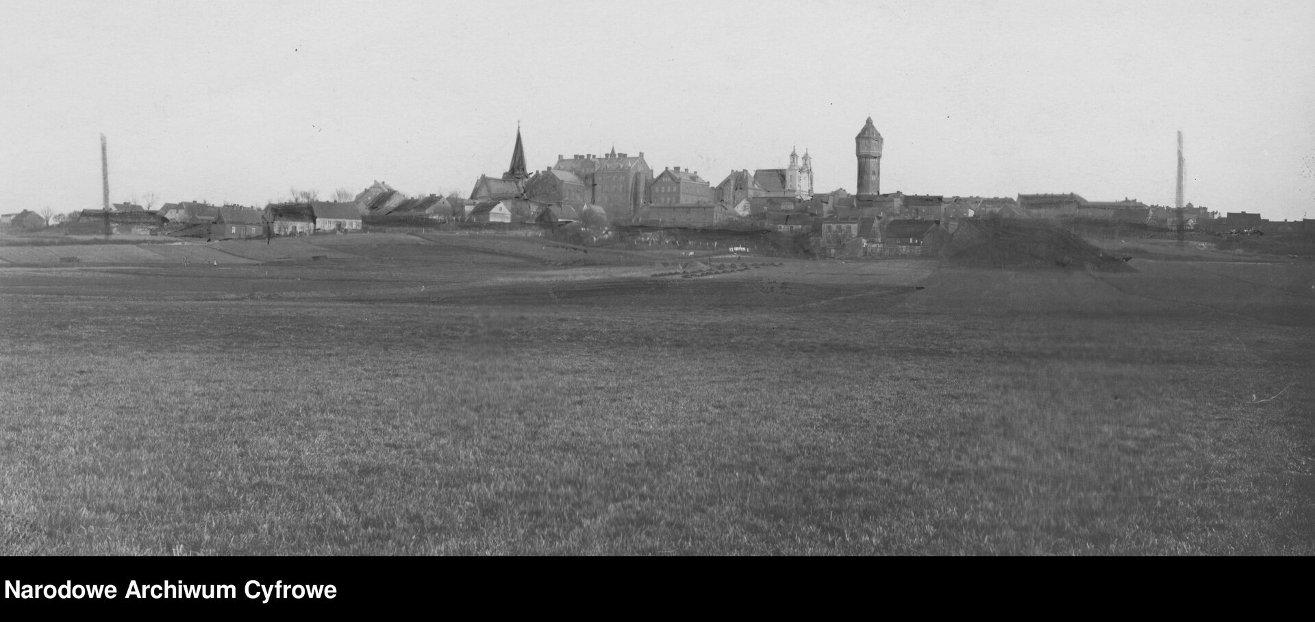 Kliknij, aby powiększyć. Zdjęcie przedstawia panoramę miasta. Na pierwszym planie znajdują się łąki i pola uprawne, dalej widać zabudowania. Nad niskimi w większości zabudowaniami górują dachy, wieża obronna (znajdująca się z prawej strony) i wieże dwóch kościołów (jedna znajduje się z lewej strony, a dwie wieże drugiego kościoła nieopodal wieży obronnej).