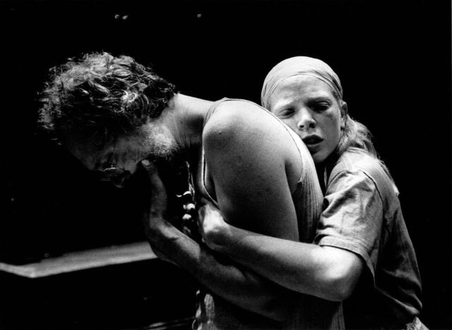 Zdjęcie przedstawia kobietę i mężczyznę. Kobieta stoi za mężczyzną przytulając się i trzymając za jego ramiona. Ma związane chustą włosy i koszulę na krótki rękaw. Mężczyzna jest w pozycji skulonej, ma opuszczoną głowę. Ubrany jest w podkoszulek, ma okulary na nosie i kręcone włosy.