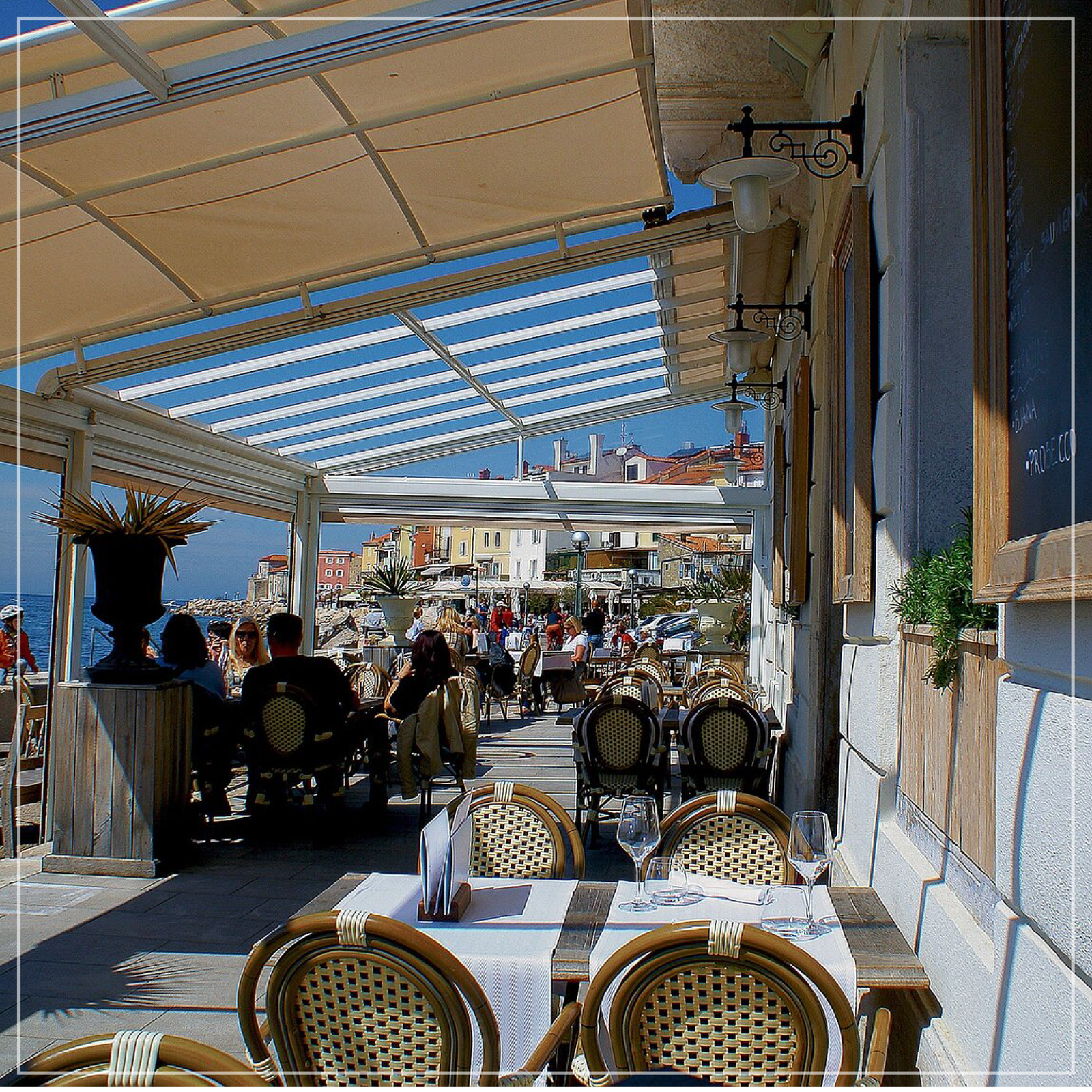 Restauracja na wybrzeżu. Na pierwszym planie puste, nakryte stoliki. Na dalszym planie ludzie siedzący przy takich samych stolikach. W oddali widać spacerujące osoby. 