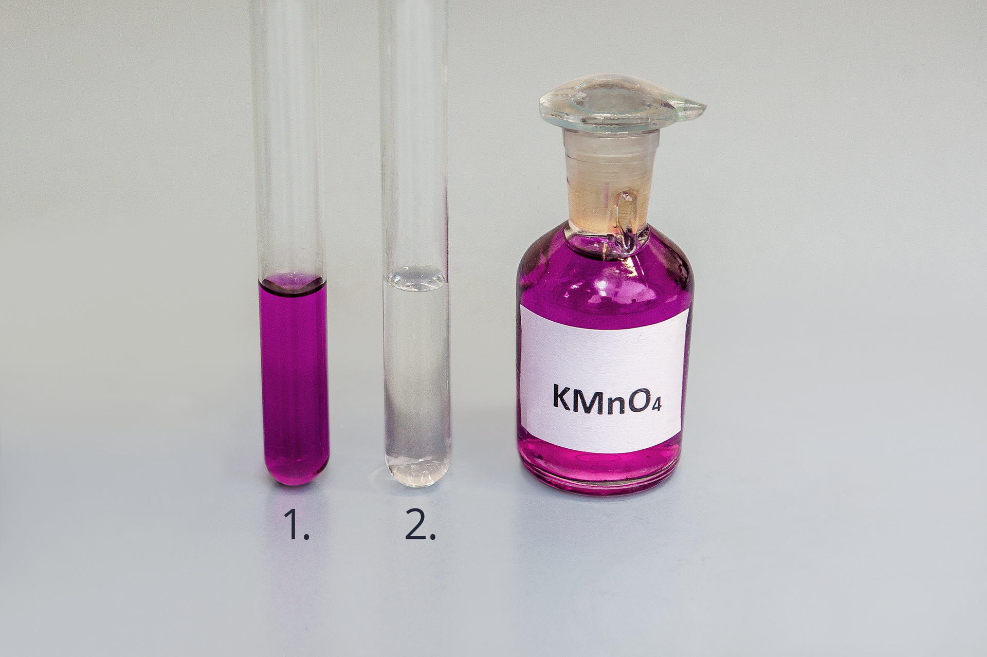 Zdjęcie przedstawia dwie probówki oraz buteleczkę z roztworem nadmanganianu potasu ka em en o cztery. W pierwszej probówce znajduje się fioletowy roztwór nadmanganianu potasu, w drugiej probówce bezbarwny roztwór. Ciecz zajmuje 1/3 objętości w obu probówkach.