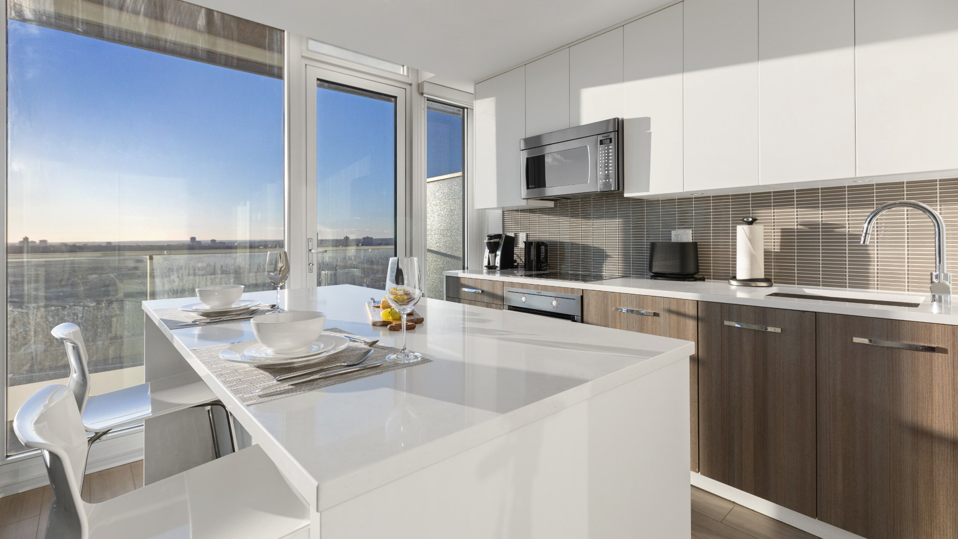 Zdjęcie przedstawia jasną kuchnię z dużym oknem. Na pierwszym planie widoczny jest stół, a przy stole dwa krzesła. Na stole znajdują się talerze, obok nich leżą sztućce. Widoczny jest także kieliszek do wina. Na drugim planie znajdują się szafki i sprzęty kuchenne. Pod blatem znajduje się piekarniki. Nad blatem widoczna jest mikrofalówka. Po prawej stronie znajduje się zlew i kran.