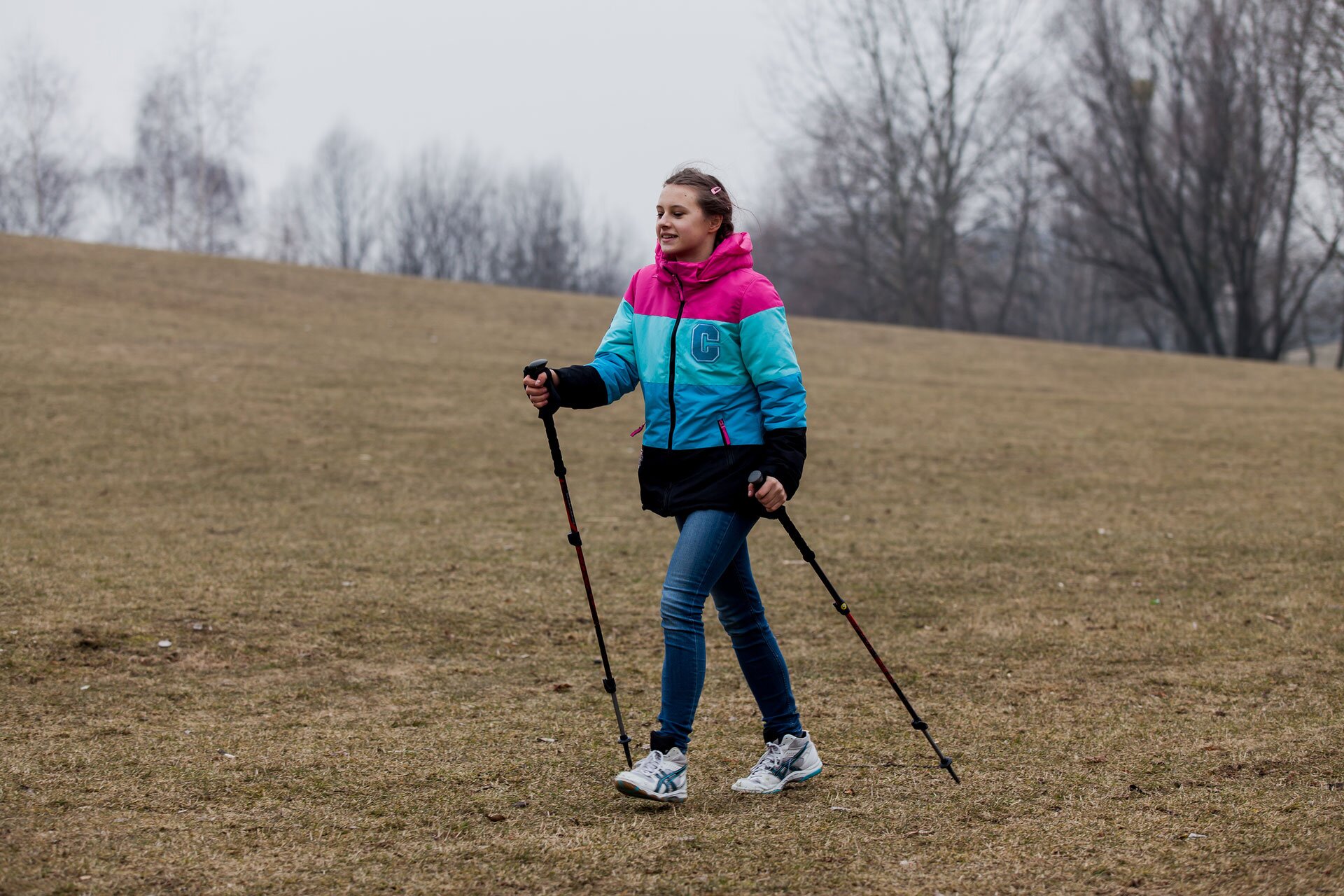 Ilustracja przedstawia dziewczynkę w ocieplanej kurtce w kolorach różowo-niebiesko-czarnych i dżinsowych wąskich spodniach. Dziewczynka idzie po niskiej, brązowej trawie i podpiera się czarnymi kijami trekkingowymi.