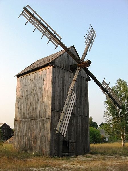 Kliknij, aby powiększyć Na zdjęciu przedstawiono drewnianą konstrukcję - wiatrak. Wyposażony jest w skrzydła, które są poruszane siłą wiatru i napędzają tą konstrukcję. Skrzydła są umieszone na samej górze, blisko dachu. Wiatrak stoi na polanie. W tle niebo.
