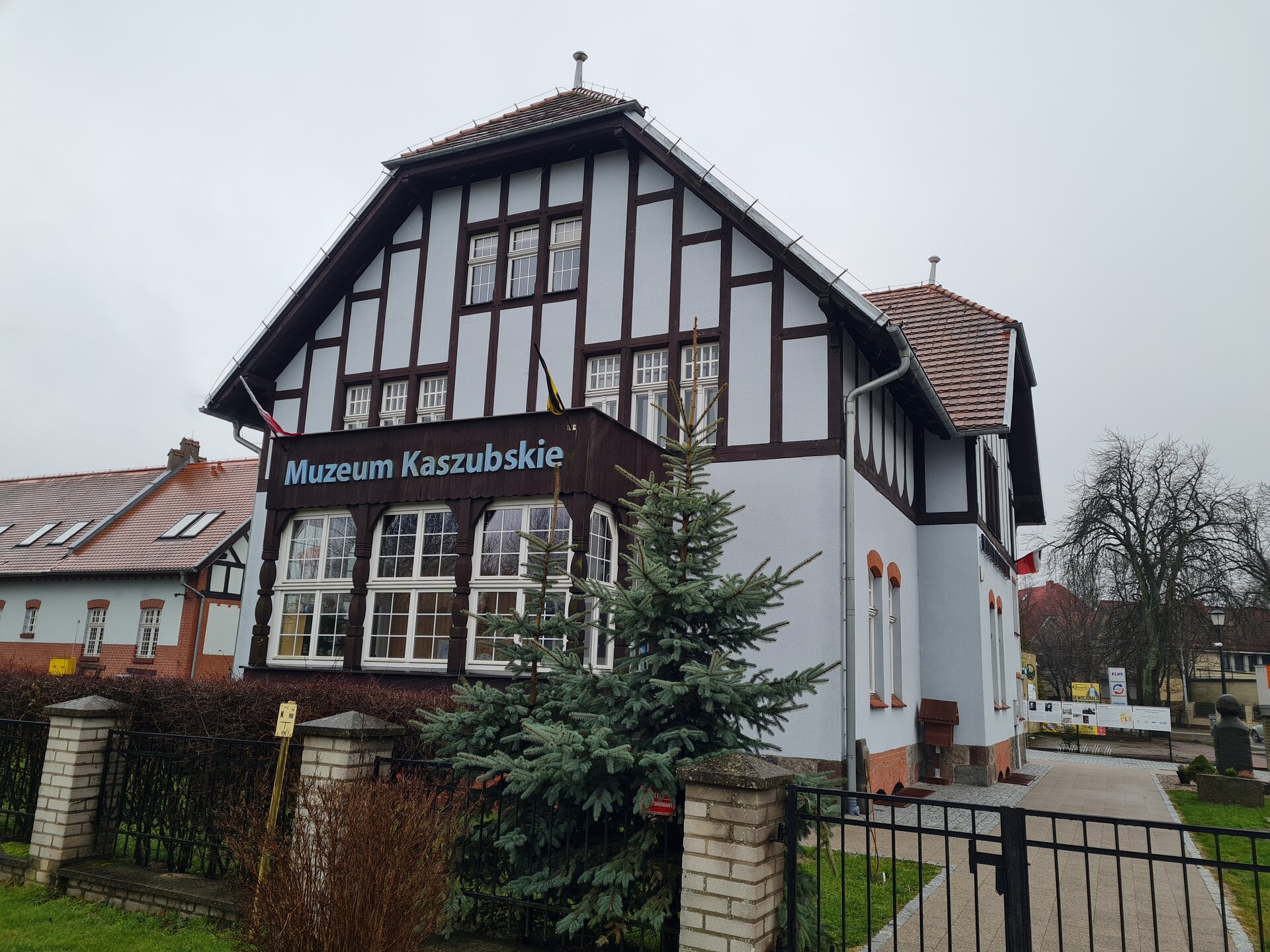 Kliknij żeby powiększyć Na zdjęciu budynek ze spadzistym dachem. Część budynku ma widoczną drewnianą konstrukcję. Umiejscowiona jest na froncie budynku. Drewniane belki są tak ułożone, że tworzą podłużne prostokąty. Na budynku widnieje napis Muzeum Kaszubskie. Budynek otoczony jest ogrodzeniem.