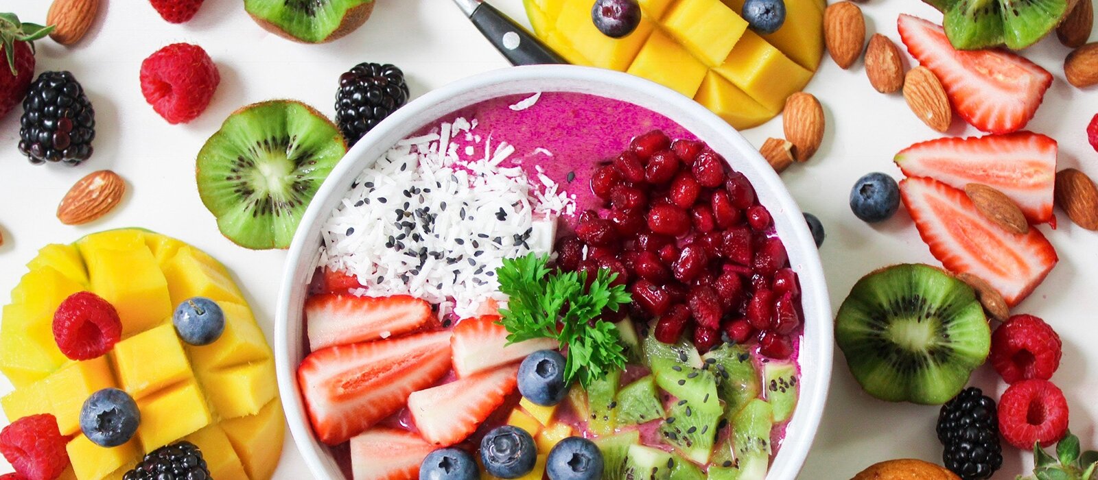 Zdjęcie przedstawia białą miskę wypełnioną różowym jogurtem oraz owocami: pokrojonymi truskawkami, kiwi, ziarnami granatu, wiórkami kokosowymi i borówkami. Dookoła miski na blacie są widoczne ułożone owoce: nadkrojona połówka mango, borówki, pokrojone kiwi i truskawki, jeżyny, maliny. Pomiędzy owocami widoczne są migdały. 