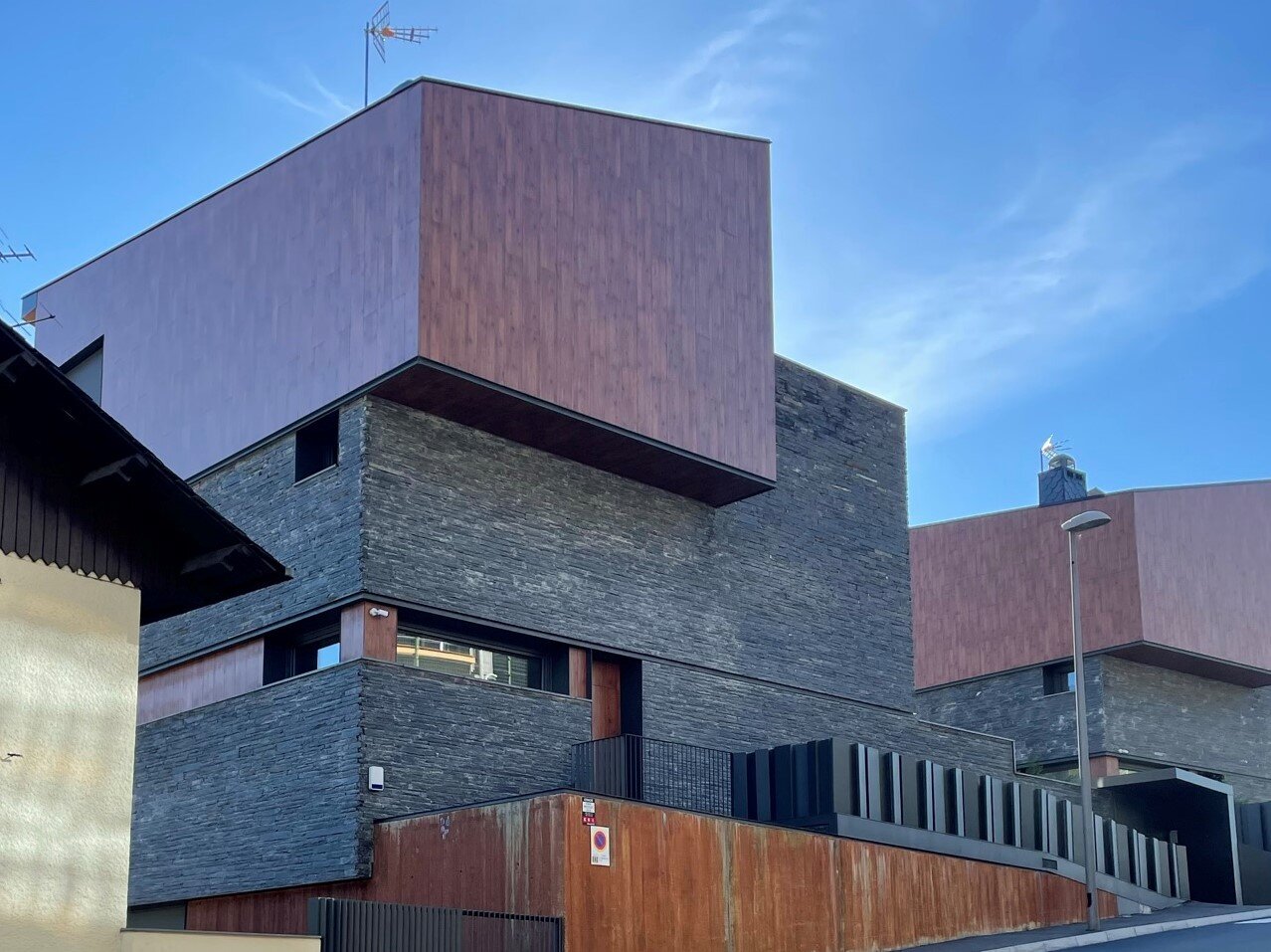 Fotografia przedstawia budynki mieszkalne w zwartej zabudowie. Na ilustracji znajdują się fasady dwóch domów usytuowanych wzdłuż ulicy. Budynki składają się z prostych brył geometrycznych: prostopadłościanów różnej wielkości, spiętrzonych nad sobą, każda pokryta inną okładziną. Bryła budynku zawiera kilka niewielkich otworów okiennych.