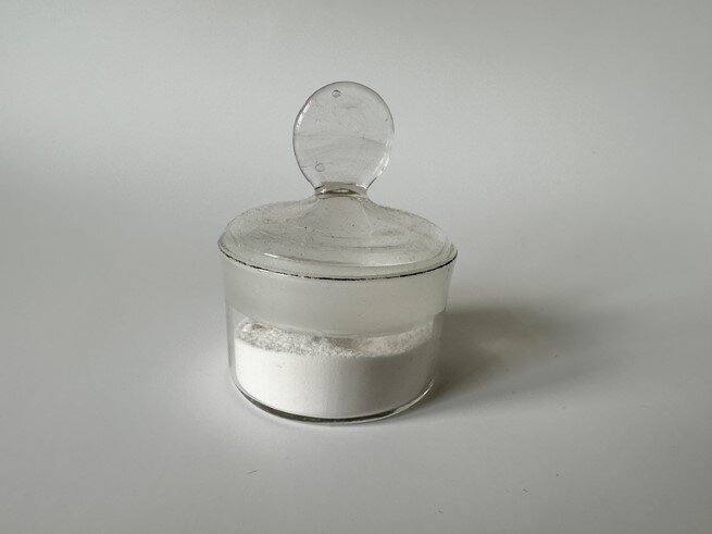 Zdjęcie przedstawia szklane naczynie z białym proszkiem - kaolinem.
