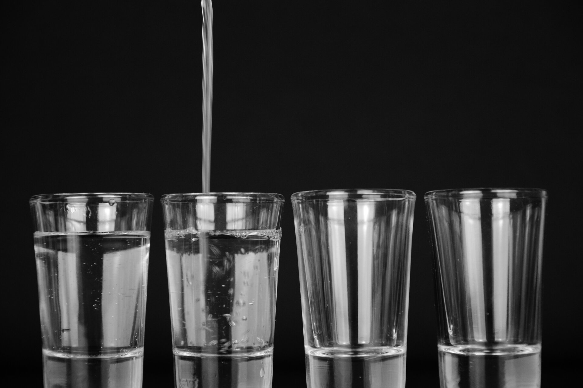 Zdjęcie przedstawia cztery szklanki stojące w jednym rzędzie. Do pierwszej szklanki nalano wodę do trzech czwartych wysokości szklanki. Do drugiej szklanki jest właśnie nalewana woda do trzech czwartych wysokości szklanki. Pozostałe dwie szklanki są puste.
