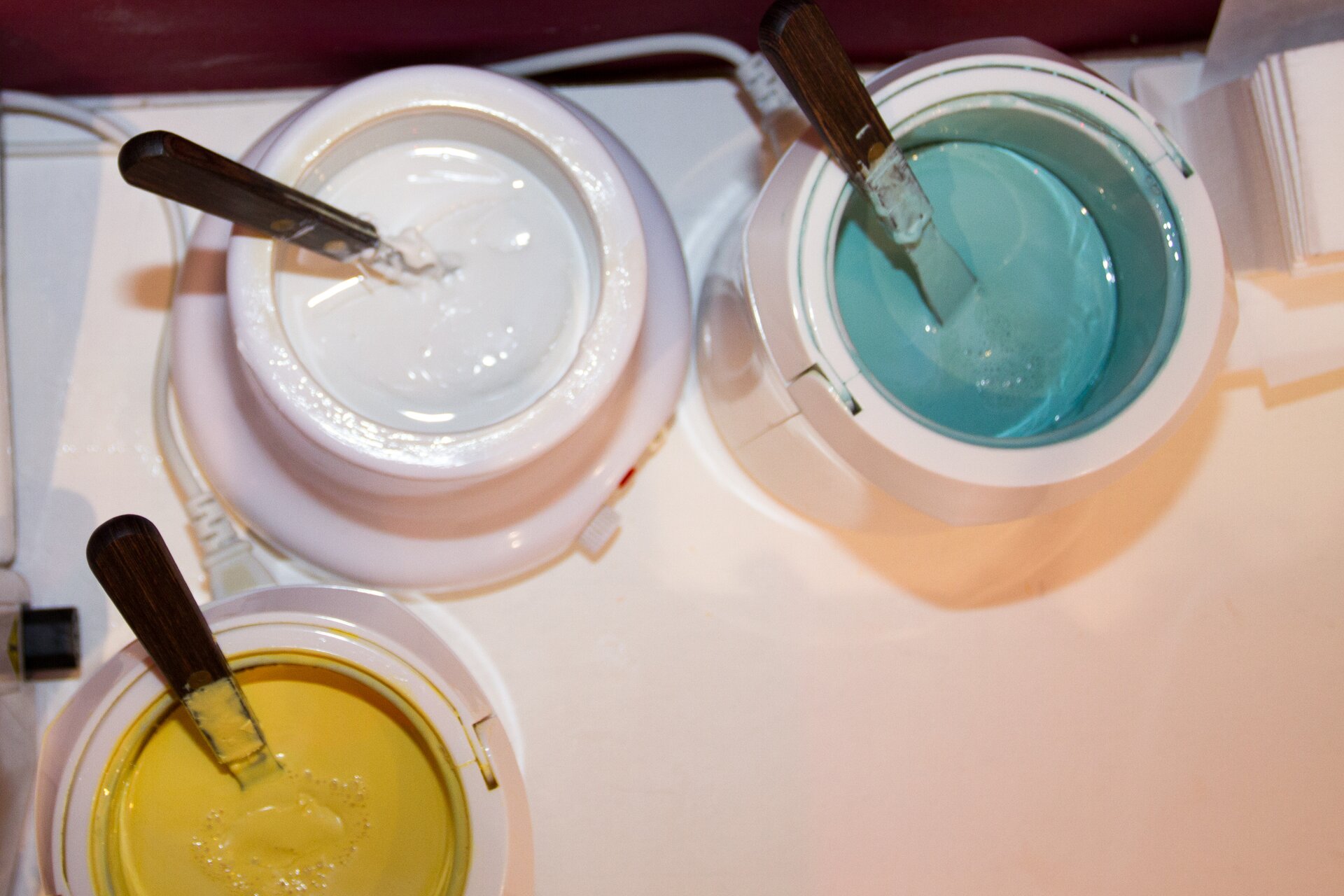 Zdjęcie przedstawia aparaturę do podgrzewania wosków depilacyjnych. Widoczne są trzy zbiorniki z woskiem o różnych kolorach. W każdym zbiorniku jest szpatułka do nakładania wosku.