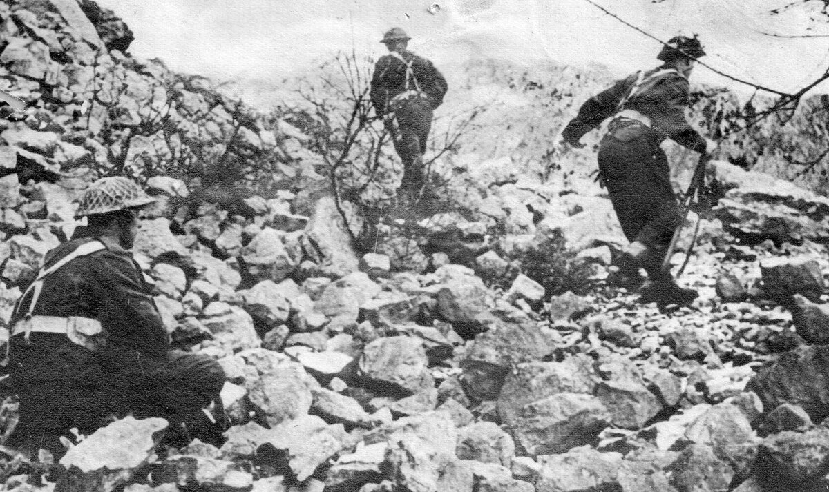 Zdjęcie przedstawia trzech żołnierzy w mundurach i kaskach. Znajdują się na gruzowisku. Żołnierz po lewej kuca, dwaj pozostali idą i są zwróceni tyłem do widza. Jeden z nich trzyma karabin.