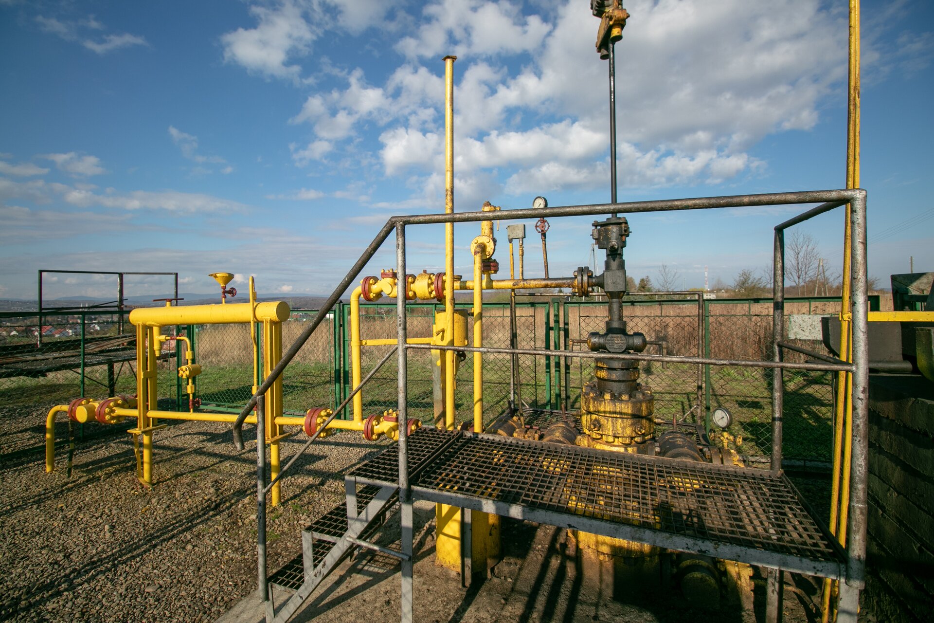 Na zdjęciu widać głowicę eksploatacyjną odwiertu pompowanego ropno‑gazowego wraz z przyległą armaturą. Głowica eksploatacyjna jest to urządzenie montowane na powierzchni terenu przy wylocie odwiertu, w celu jego zamknięcia. Na zdjęciu widać, że głowica to stalowe urządzenie pomalowane na kolor żółty. Na górze urządzenia znajdują się czarne zacieki ropy. Podstawę urządzenia stanowi walcowaty kształt czyli kołnierz. Na nim znajduje się prwenter czyli urządzenie zabezpieczające. Prewenter jest w kształcie krzyża. Na prewentrze znajduje się trojak czyli rura w kształcie litery T umożliwiająca rozgałęzienie. Na górze urządzenia znajduje się laska pompowa w kształcie walcowatego pionowego pręta. Do końcówki trojaka, która jest skierowana w lewą stronę przyłączona jest armatura rurowa wraz z różnym typem zaworów. Przed głowicą znajduje się stalowy pomost. Cały układ zlokalizowany jest w wygrodzonym terenie. Przestrzeń oddzielona jest zieloną metalową siatką. Za siatką widać ciągnące się pole, zaś z lewej strony widać w oddali zabudowania mieszkalne.