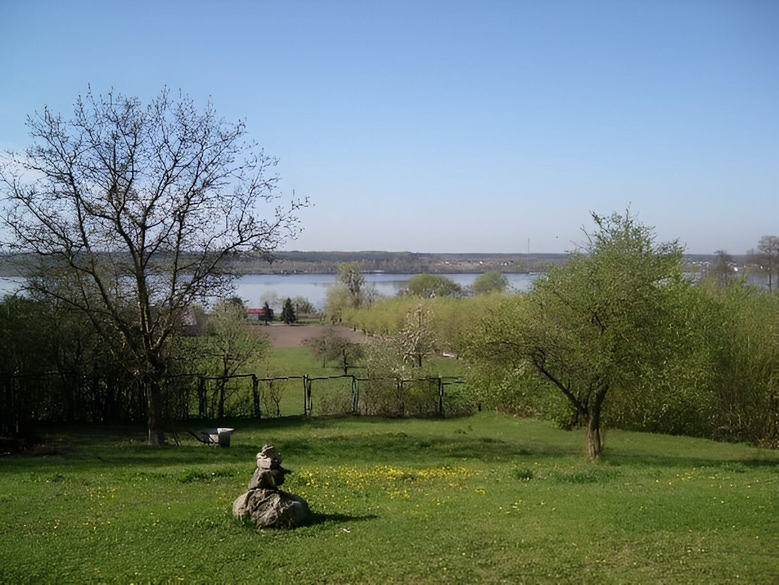 Fotografia przedstawia łąkę, na niej po prawej i lewej stronie kępy drzew. Na pierwszym planie kamienie ułożone jeden na drugim. Za nimi płot. W tle rozległa rzeka.