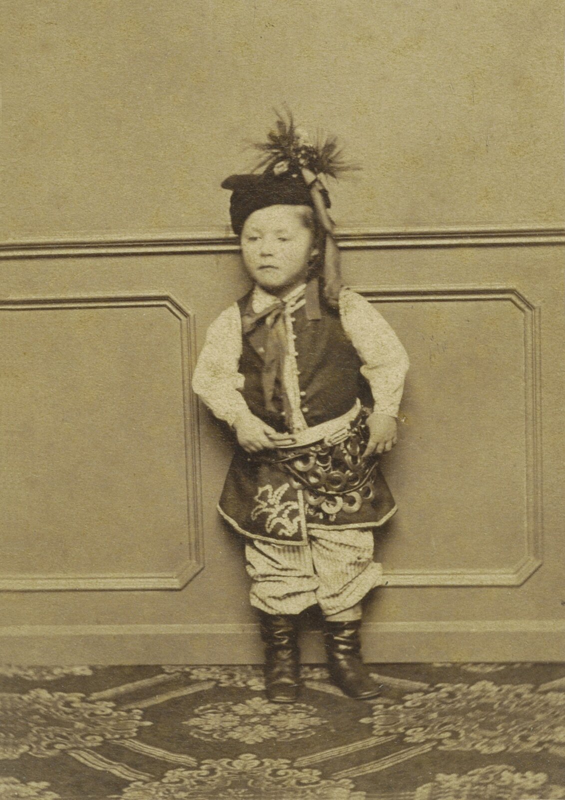 Zdjęcie czarno‑białe. Portret dziecka w stroju krakowskim, ok. 1865. Chłopczyk w białej koszuli, w ozdobnym długim kaftanie,  pasiastych spodenkach. Pod szyją ma zawiązaną wstążkę. Na głowie czapkę z kokardą i piórami.