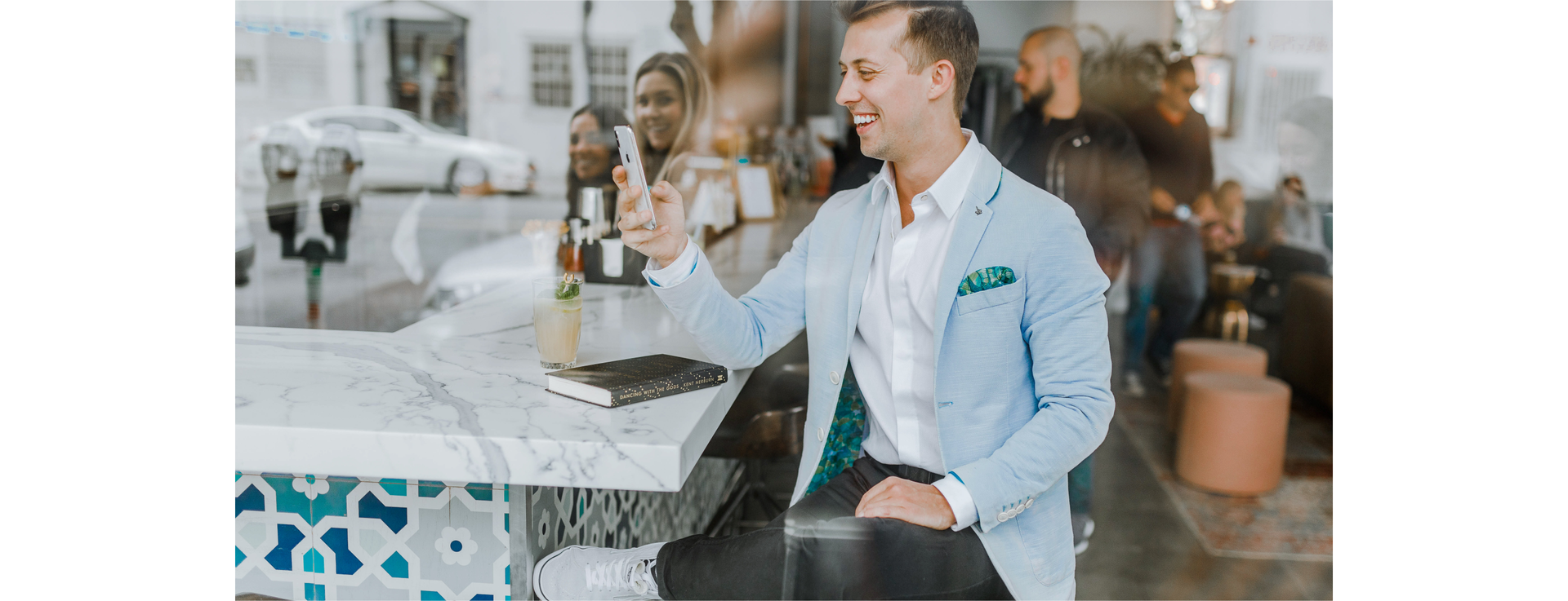 Zdjęcie przedstawia elegancko ubranego, uśmiechniętego mężczyznę. Mężczyzna siedzi przy barze i spogląda w telefon komórkowy. Na blacie leży jego notes.