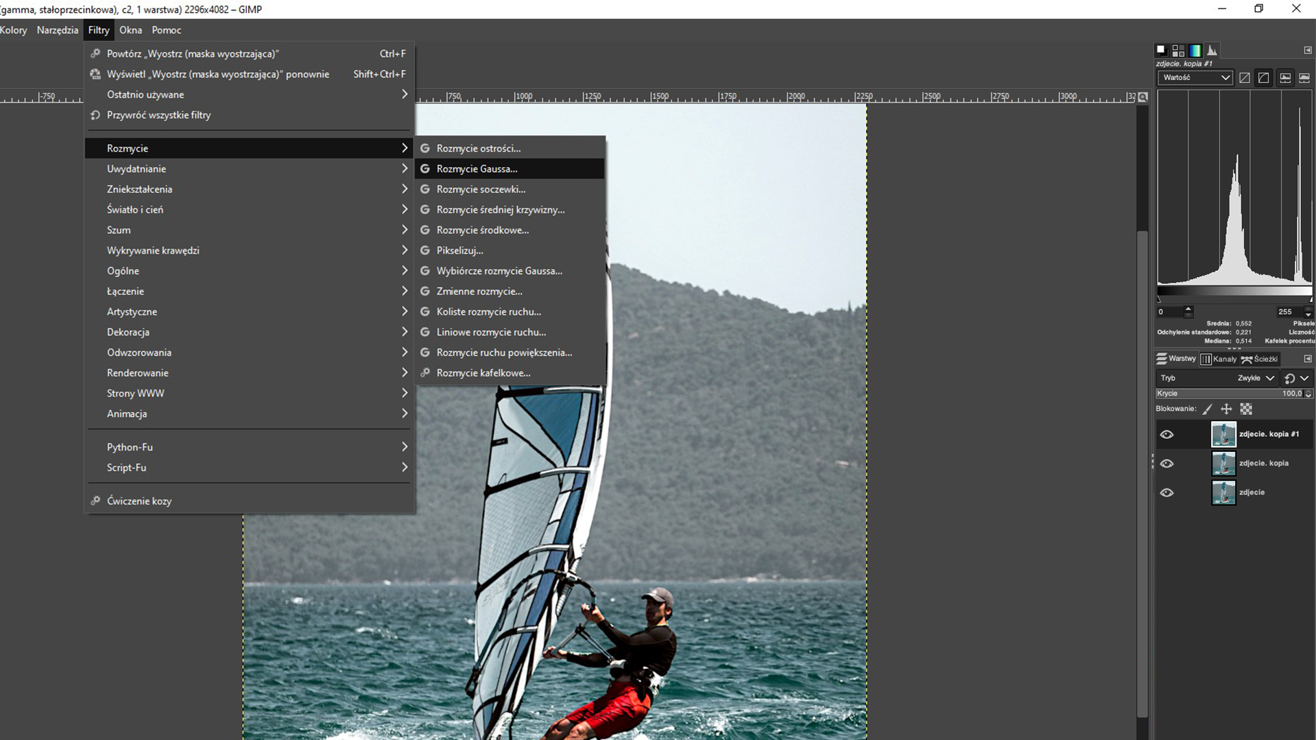Ilustracja przedstawia okno programu. W obszarze roboczym jest zdjęcie mężczyzny na windsurfingu. Z menu wybrano zakładkę: Filtry, tam z listy wybrano: Rozmycie, a następnie Rozmycie Gaussa. Po prawej stronie jest panel dotyczący warstw. Tam zdjecie.kopia #1, zdjęcie.kopia, zdjecie. Zaznaczono zdjecie.kopia #1.  