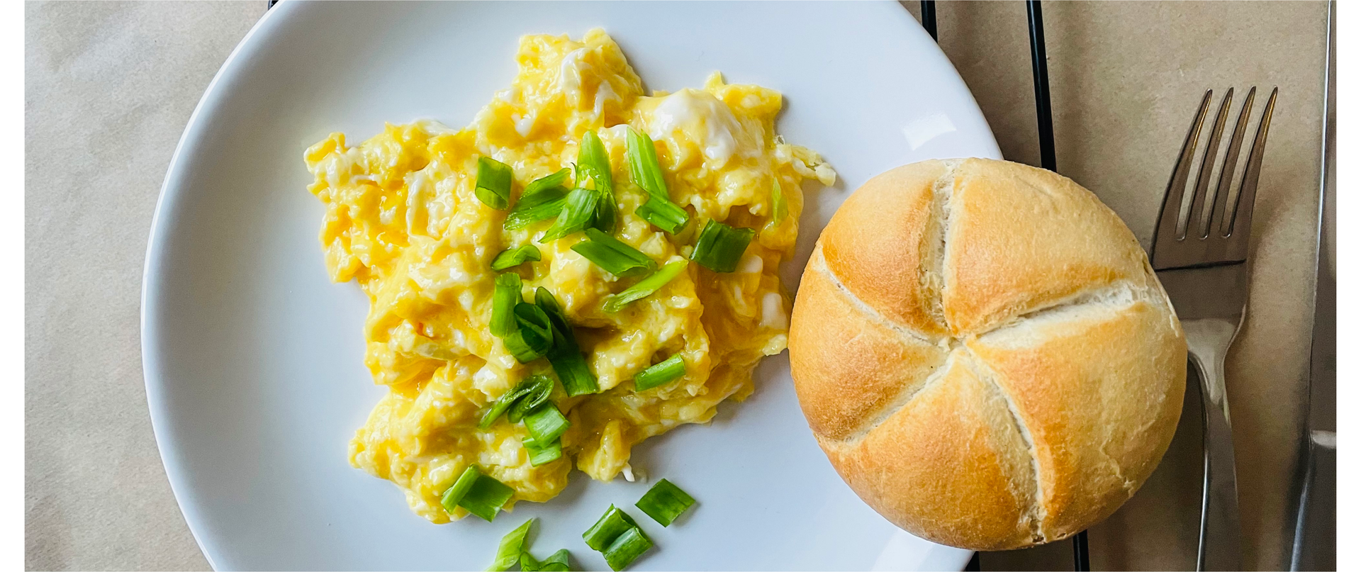 Zdjęcie przedstawia jajecznicę ze szczypiorkiem na talerzu. Obok jajecznicy leży bułka kajzerka. Po prawej stronie widoczne są sztućce.