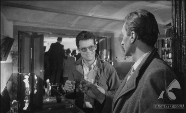 Zdjęcie przedstawia wnętrze baru. Przy długiej barowej ladzie stoi dwóch mężczyzn, którzy patrzą na siebie. Wznoszą toast, trzymając w dłoniach kieliszki z wódką.