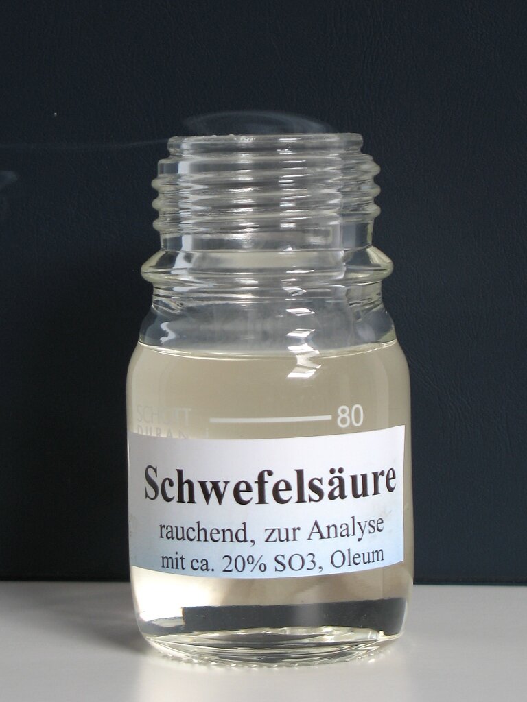 Ilustracja przedstawia szklany słoiczek zawierający przeźroczystą substancję, słoik jest pełny. Nie jest zakręcony. Na słoiku widnieje biała etykieta zawierająca czarny napis Schwefelsäure, rauchend, zur analyse mit ca. 20 % es o trzy, Oleum.